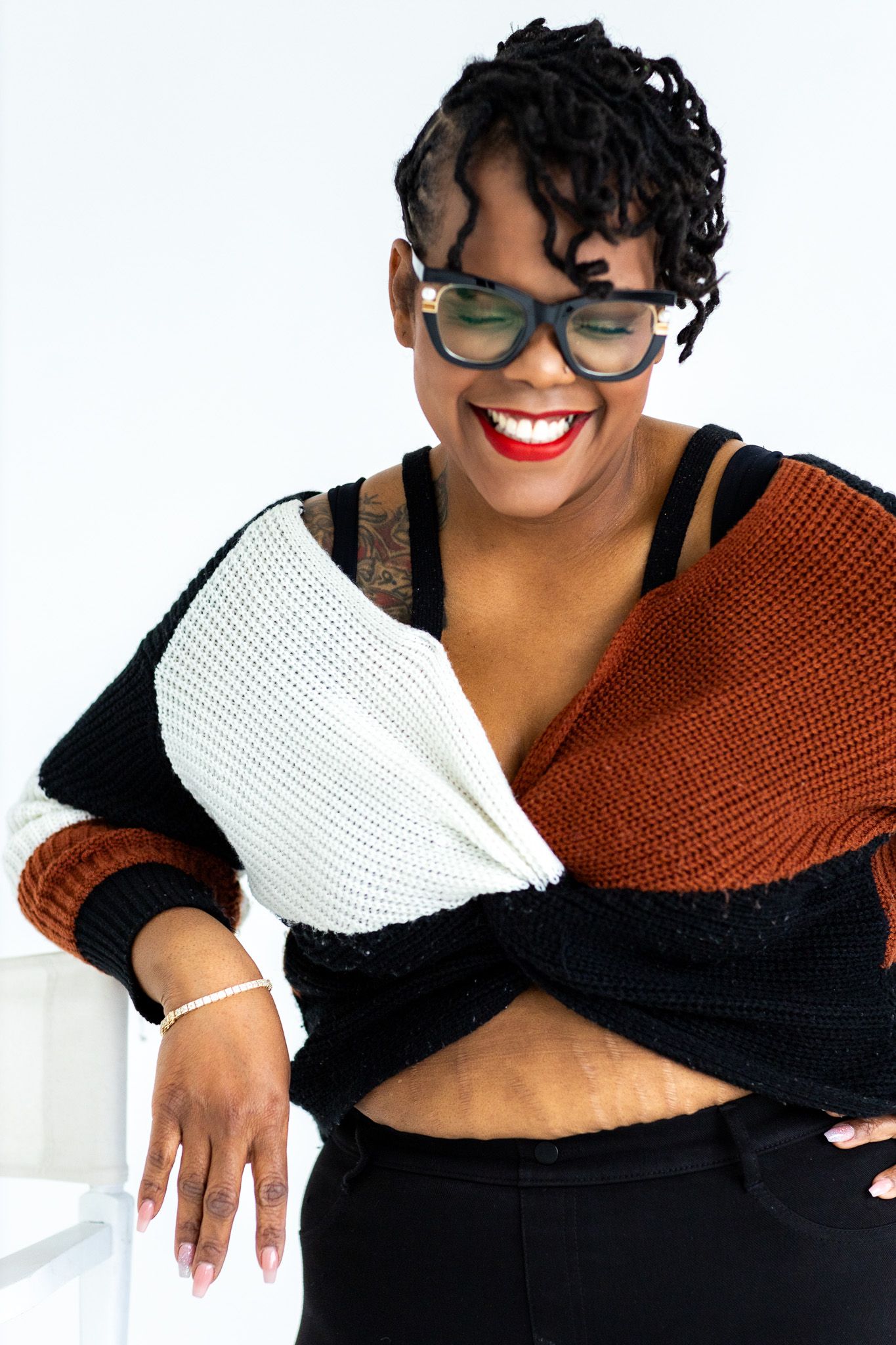 Smiling Black woman in colorblock knit sweater with glasses, red lips, and braids poses against white background.