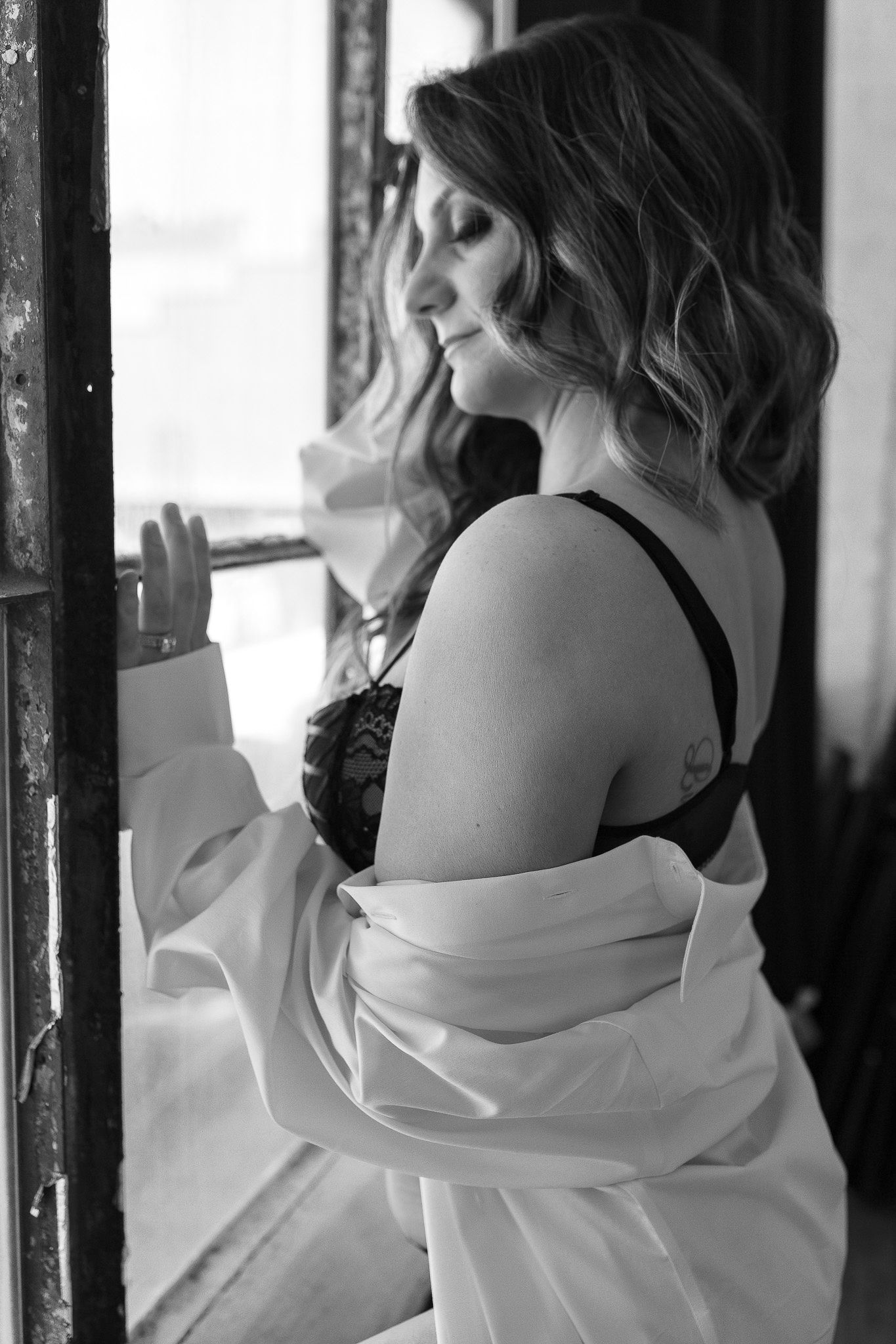 Black and white boudoir photo of a woman in black lace bra with white shirt off her shoulders by a window.