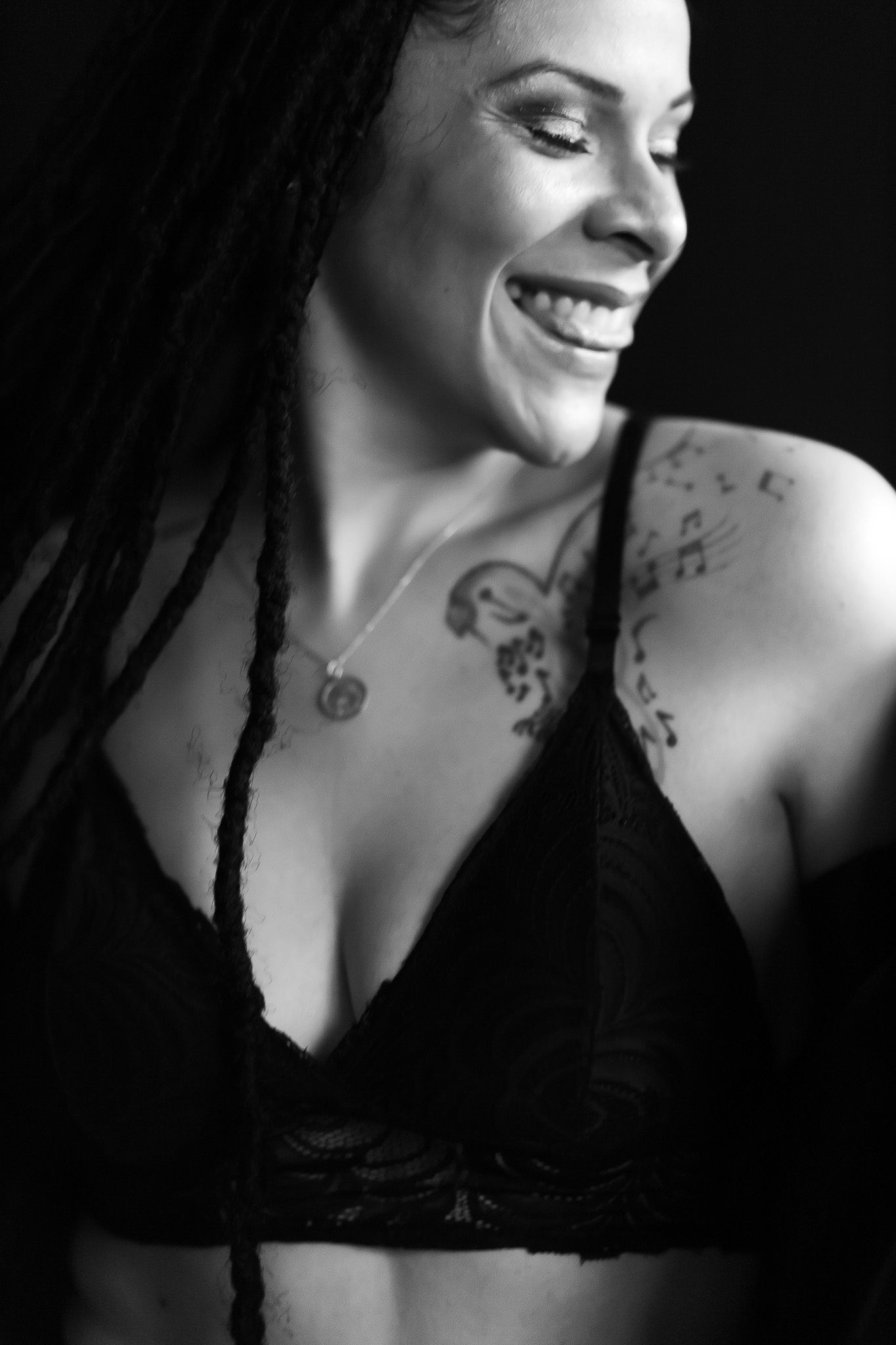 Close-up black and white portrait of a laughing woman with braids and musical note chest tattoos in a lace bra.