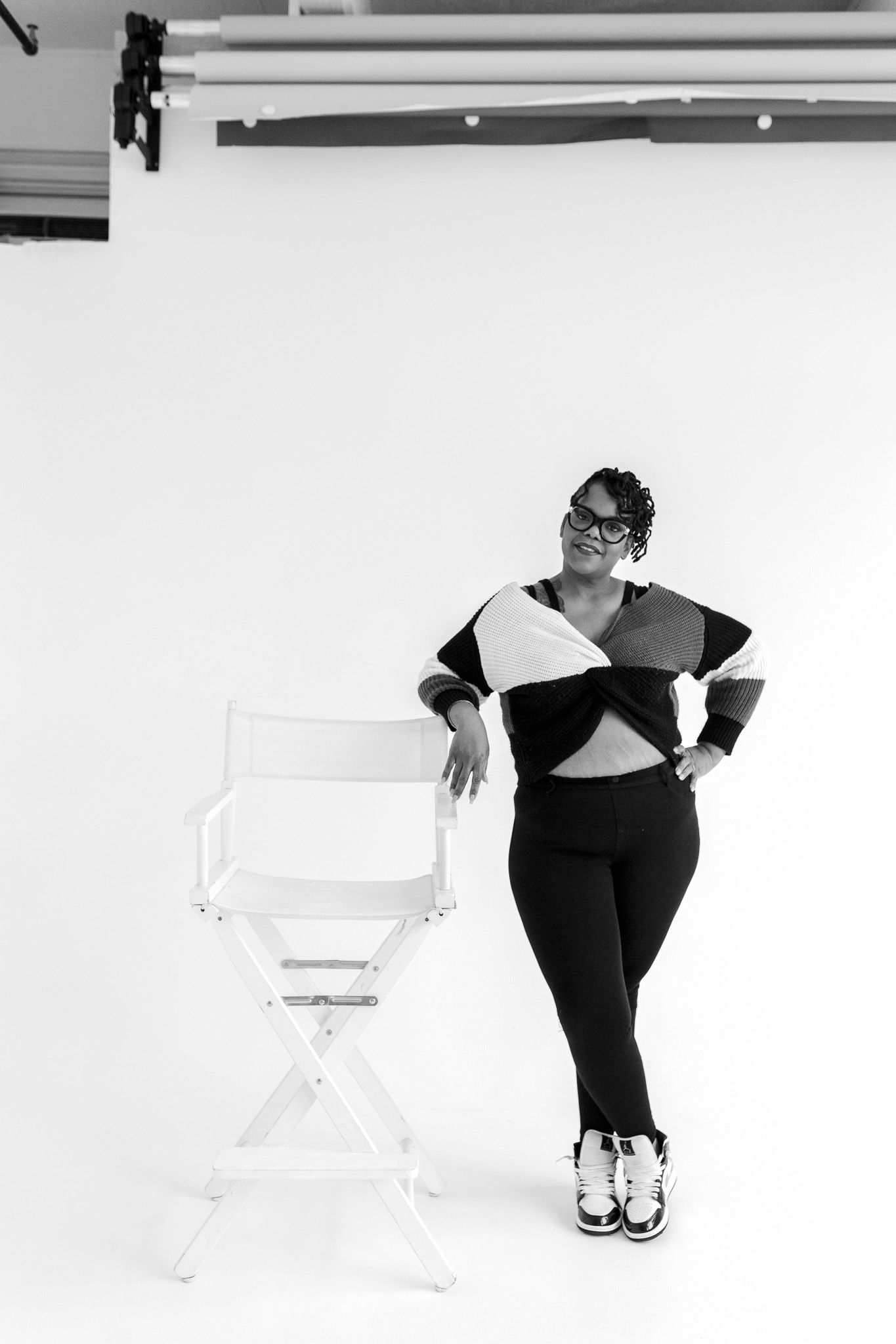 Black and white photo of a woman in stylish outfit leaning on a director's chair in a photography studio.