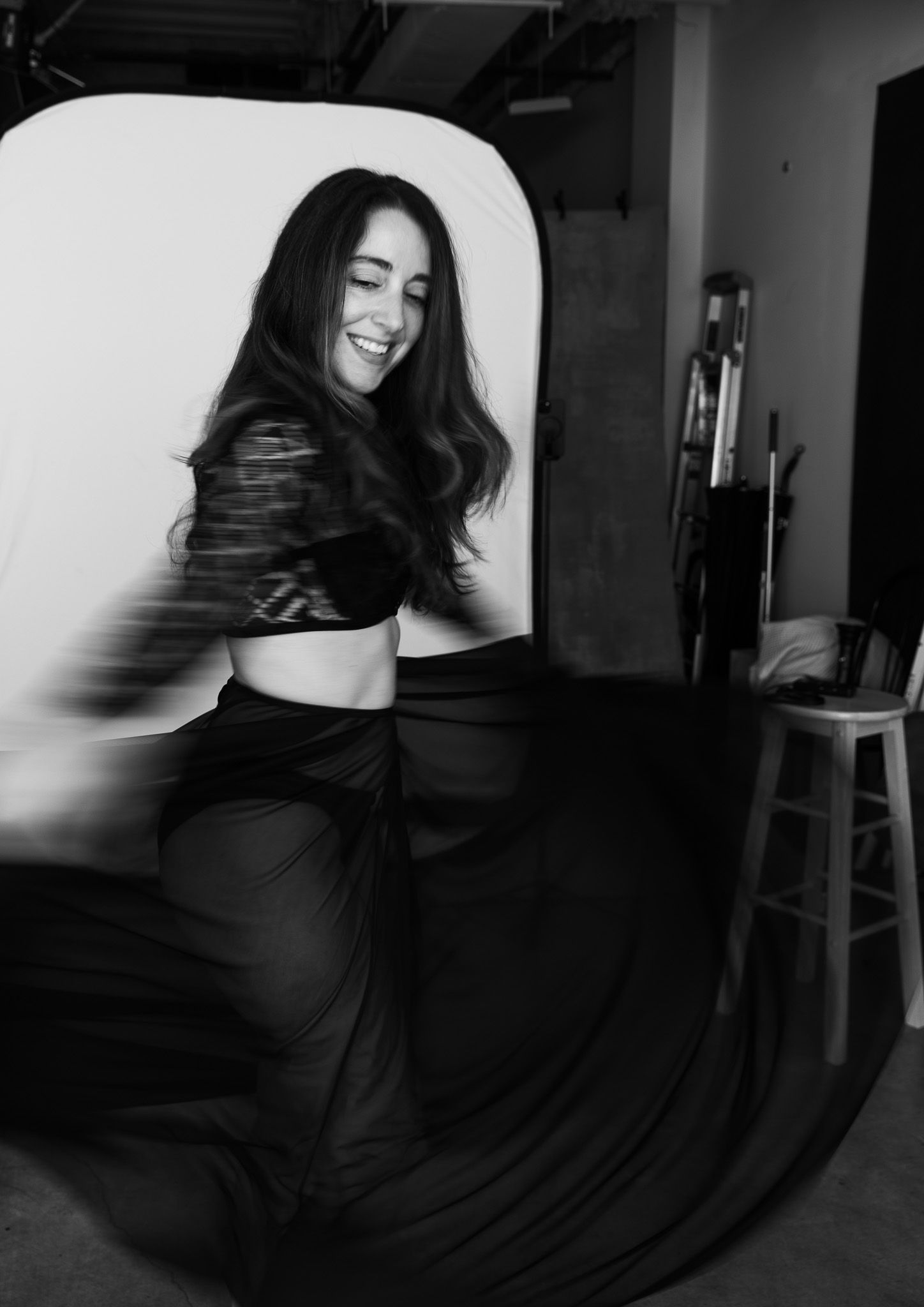 Black and white photo of a joyful woman laughing while dancing in a flowing black skirt in a studio.