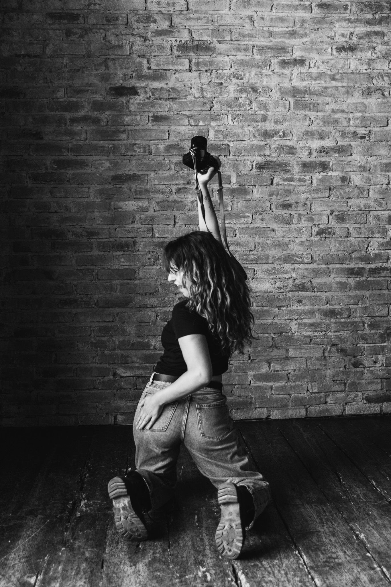 Black and white photo of a woman in jeans kneeling against a brick wall holding a camera strap.
