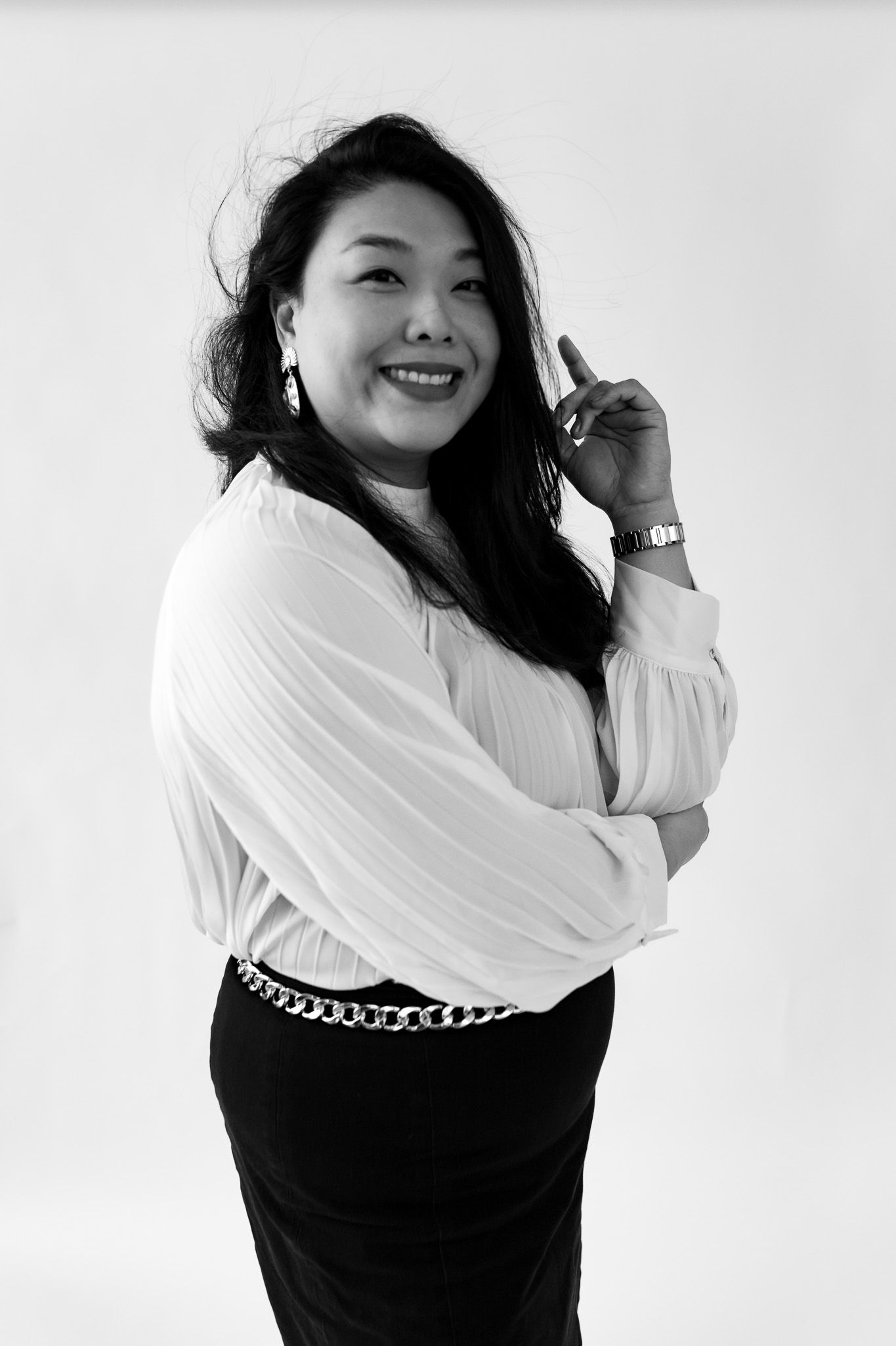 Black and white photo of a smiling Asian woman in white blouse and black skirt with chain belt, finger raised.