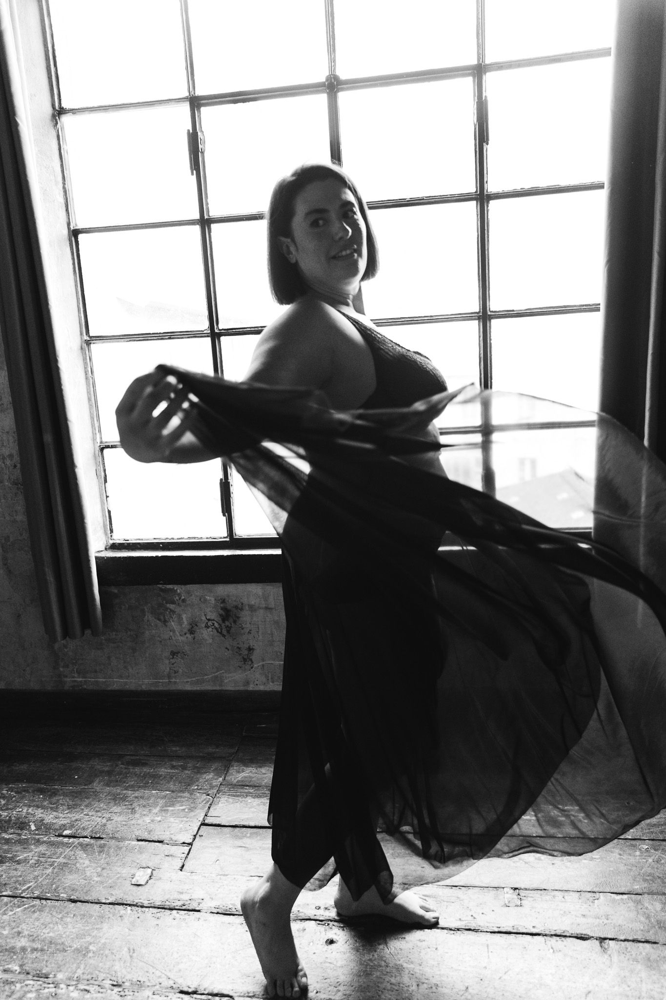 Black and white photo of a woman dancing gracefully in flowing dark fabric near a large industrial window.