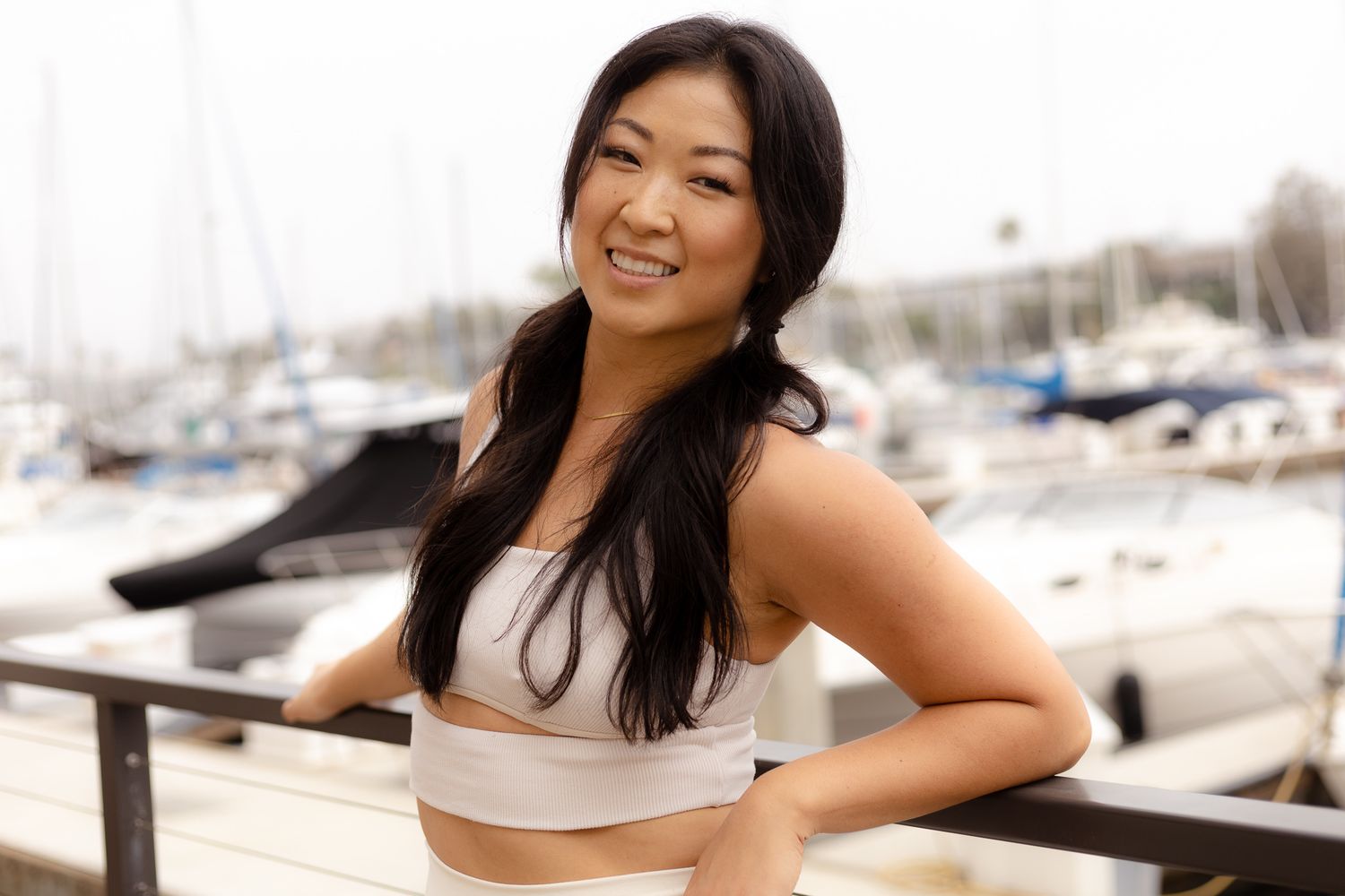Smiling young woman in white crop top posing at a marina with sailboats in the background.