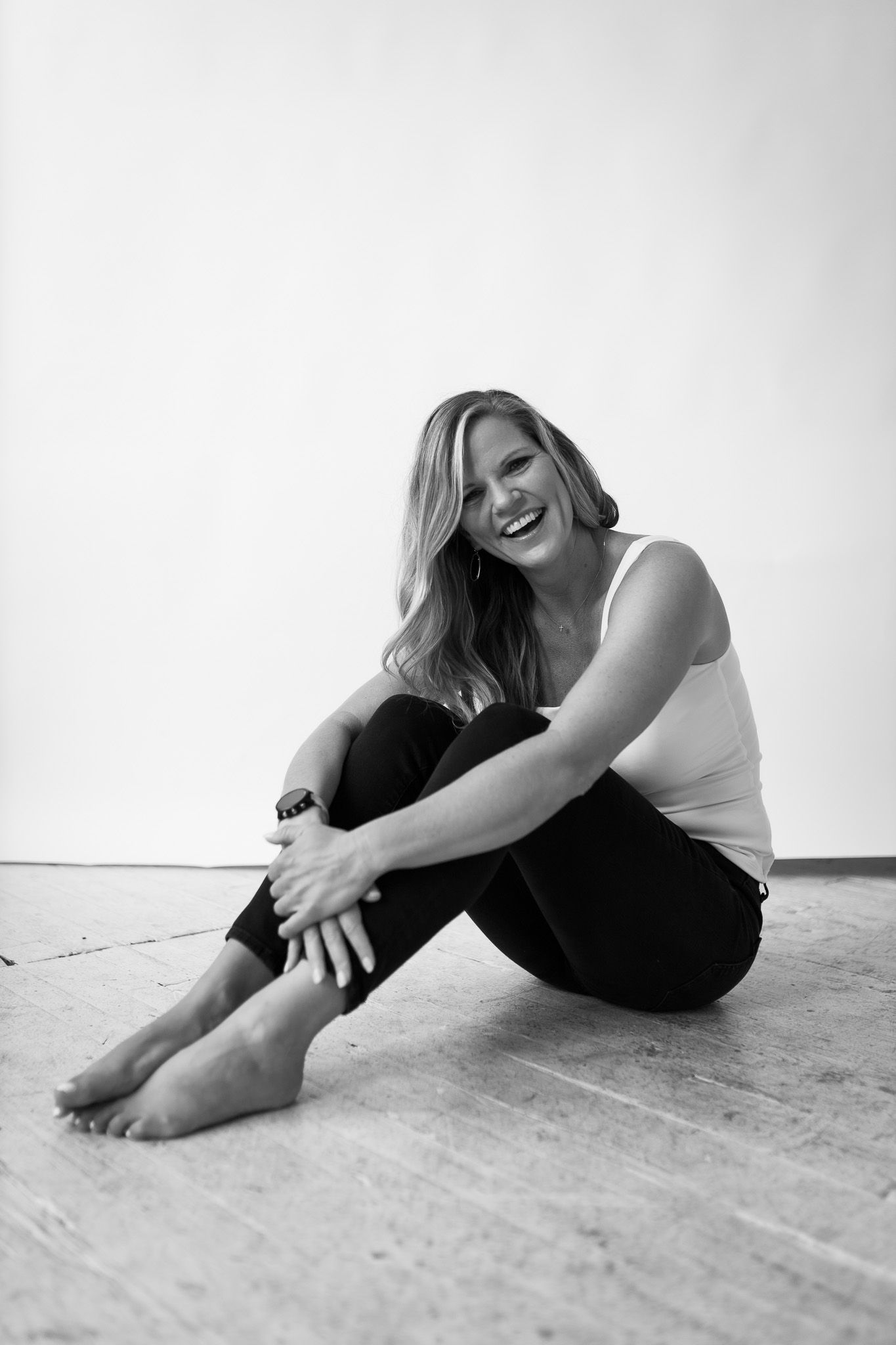 Smiling blonde woman sitting barefoot on wooden floor in white tank top and black jeans, black and white photo.