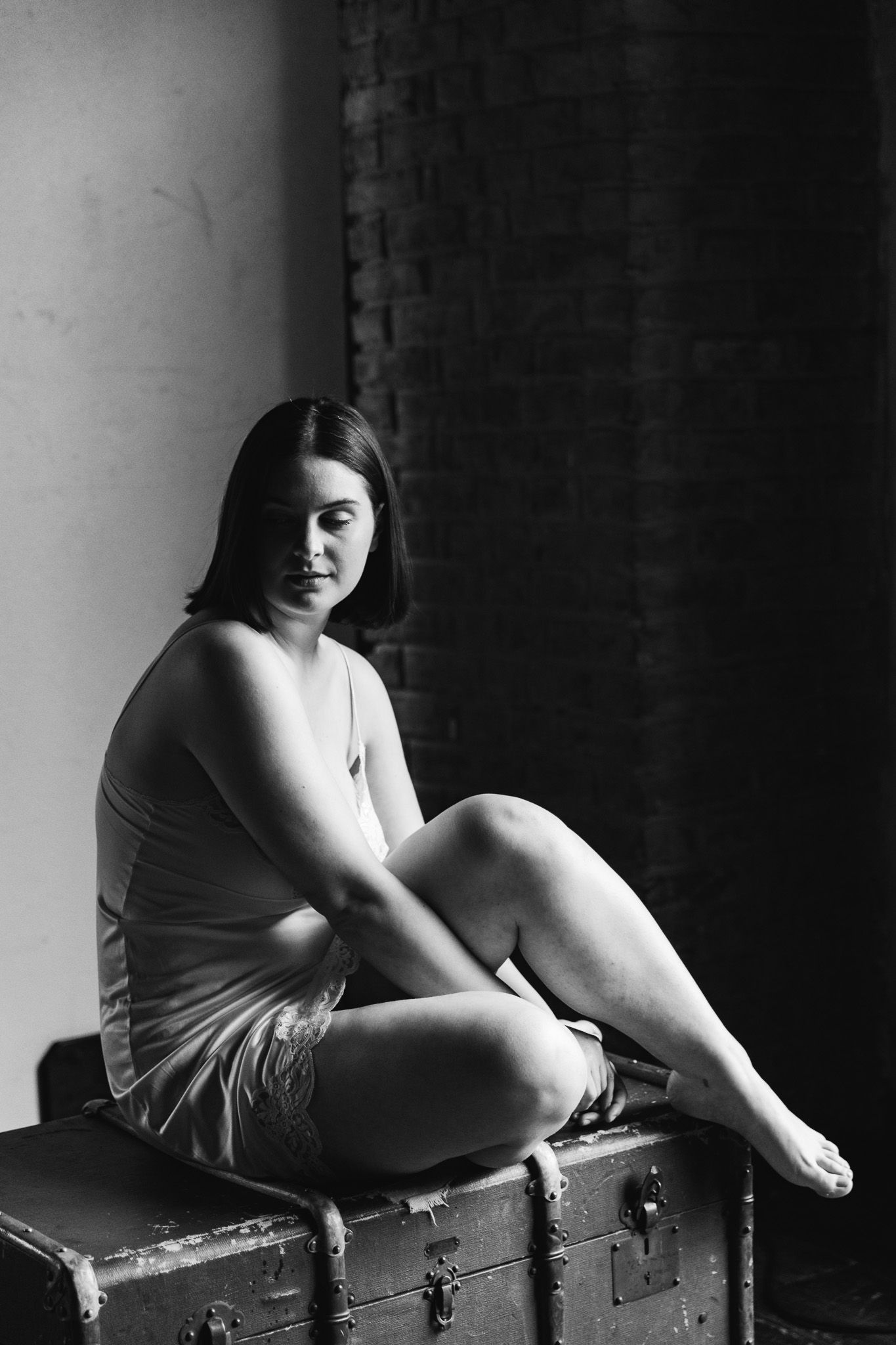 Moody black and white photo of a woman in a tank dress seated on an antique trunk near brick wall.
