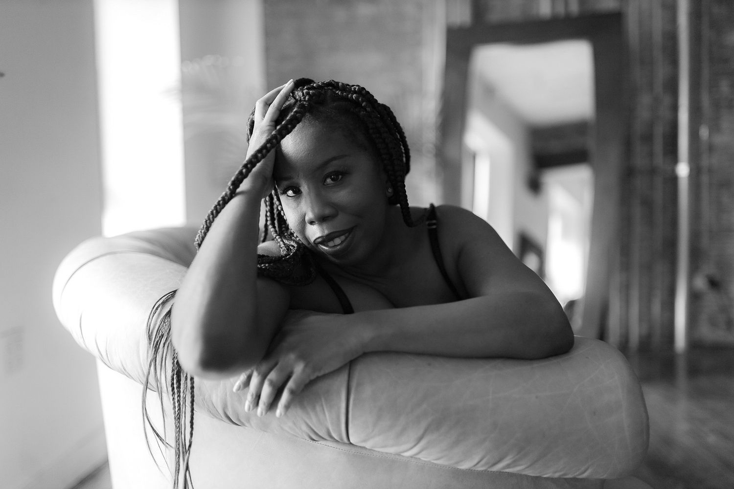 Black and white portrait of a smiling woman with braids leaning on a chair.