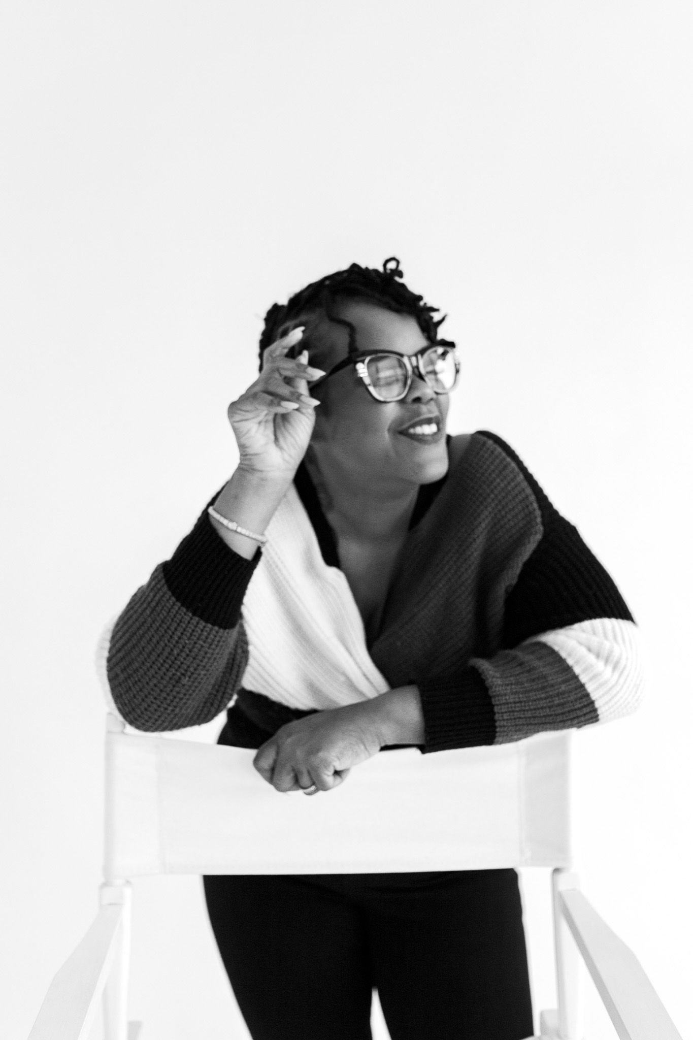 Black and white portrait of a smiling woman adjusting her glasses while leaning on a white chair.