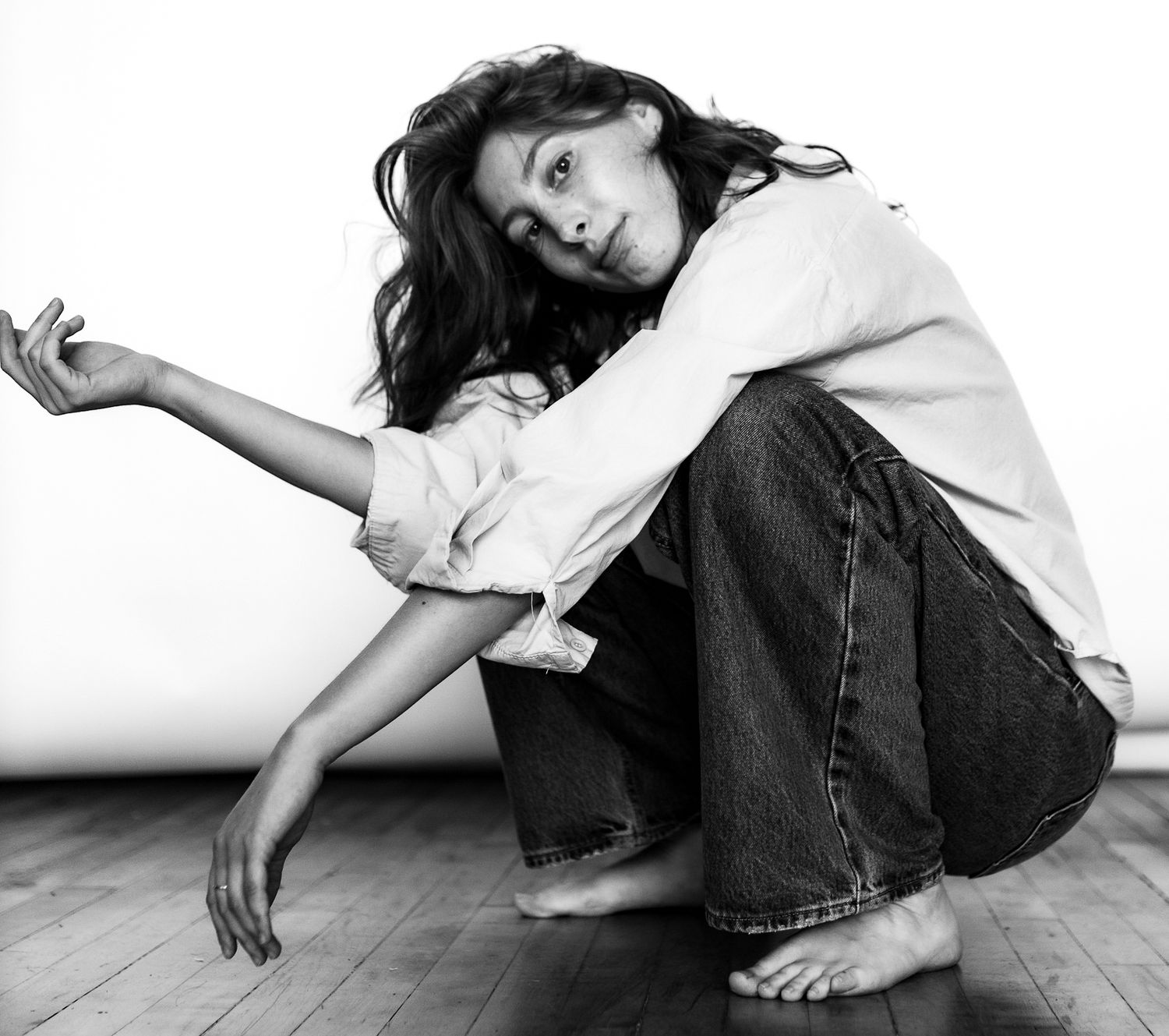 Woman in white shirt and wide-leg jeans crouches on floor in expressive black and white photo.