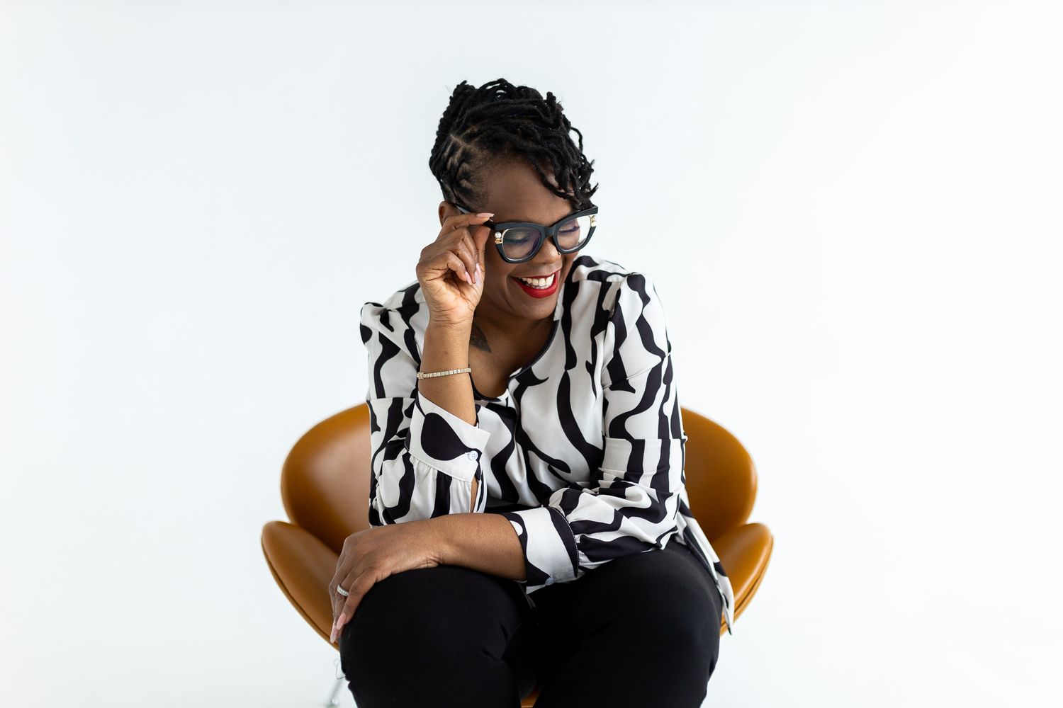 Confident Black woman in zebra print blouse adjusts glasses while seated in brown leather chair, smiling.
