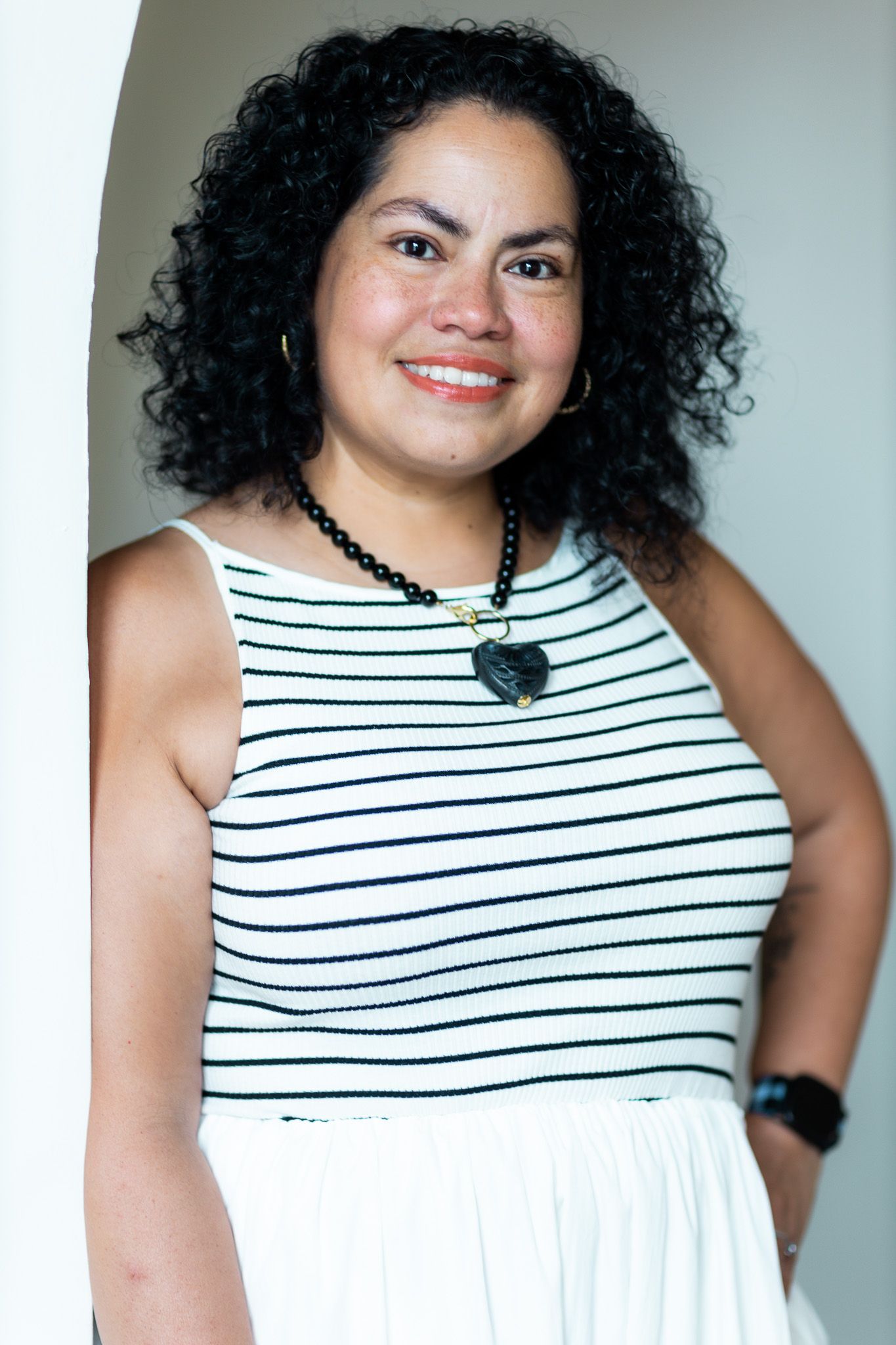Smiling woman with curly black hair wearing striped top and heart necklace leaning against white pillar.