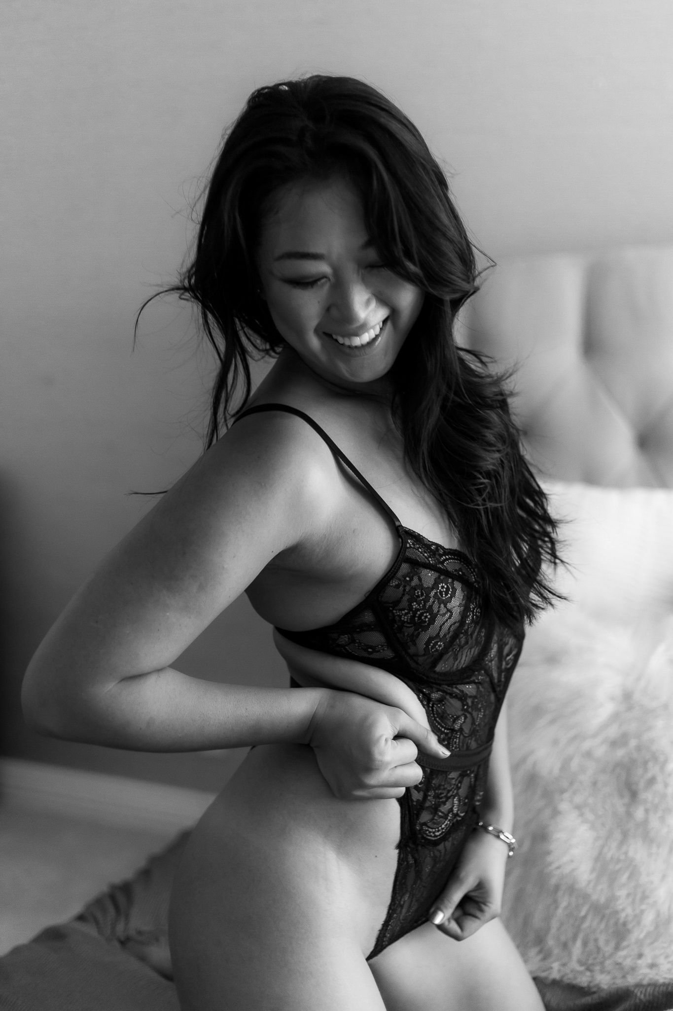 Black and white boudoir photo of smiling woman in lace lingerie posing on bed.