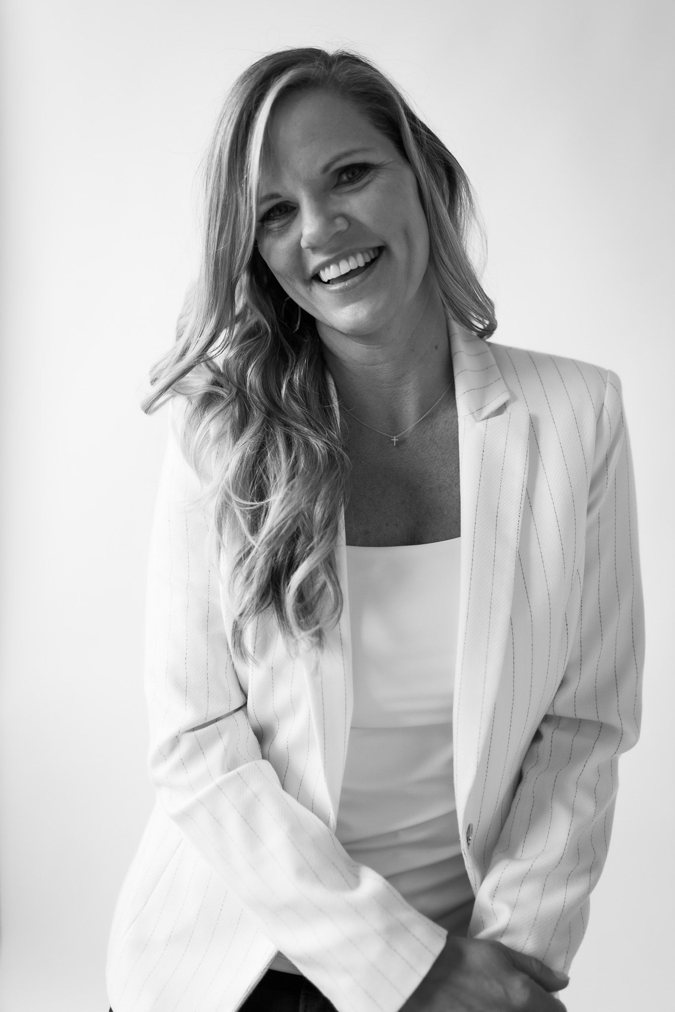 Smiling woman with long blonde hair wearing a white blazer and top, posed confidently in a black and white studio portrait.