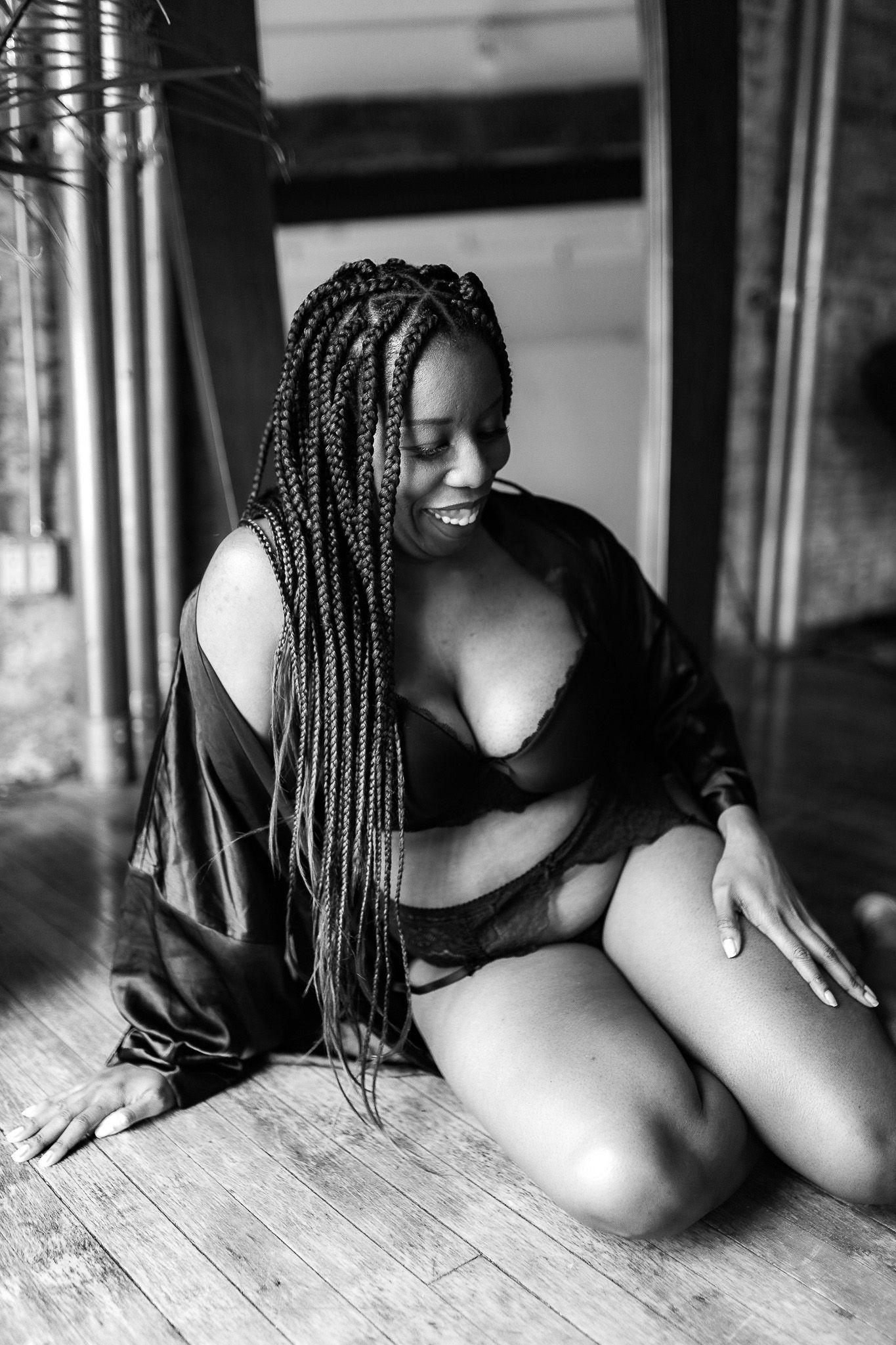 Black woman with long box braids poses on hardwood floor in black lingerie and open robe, elegant black and white boudoir portrait.