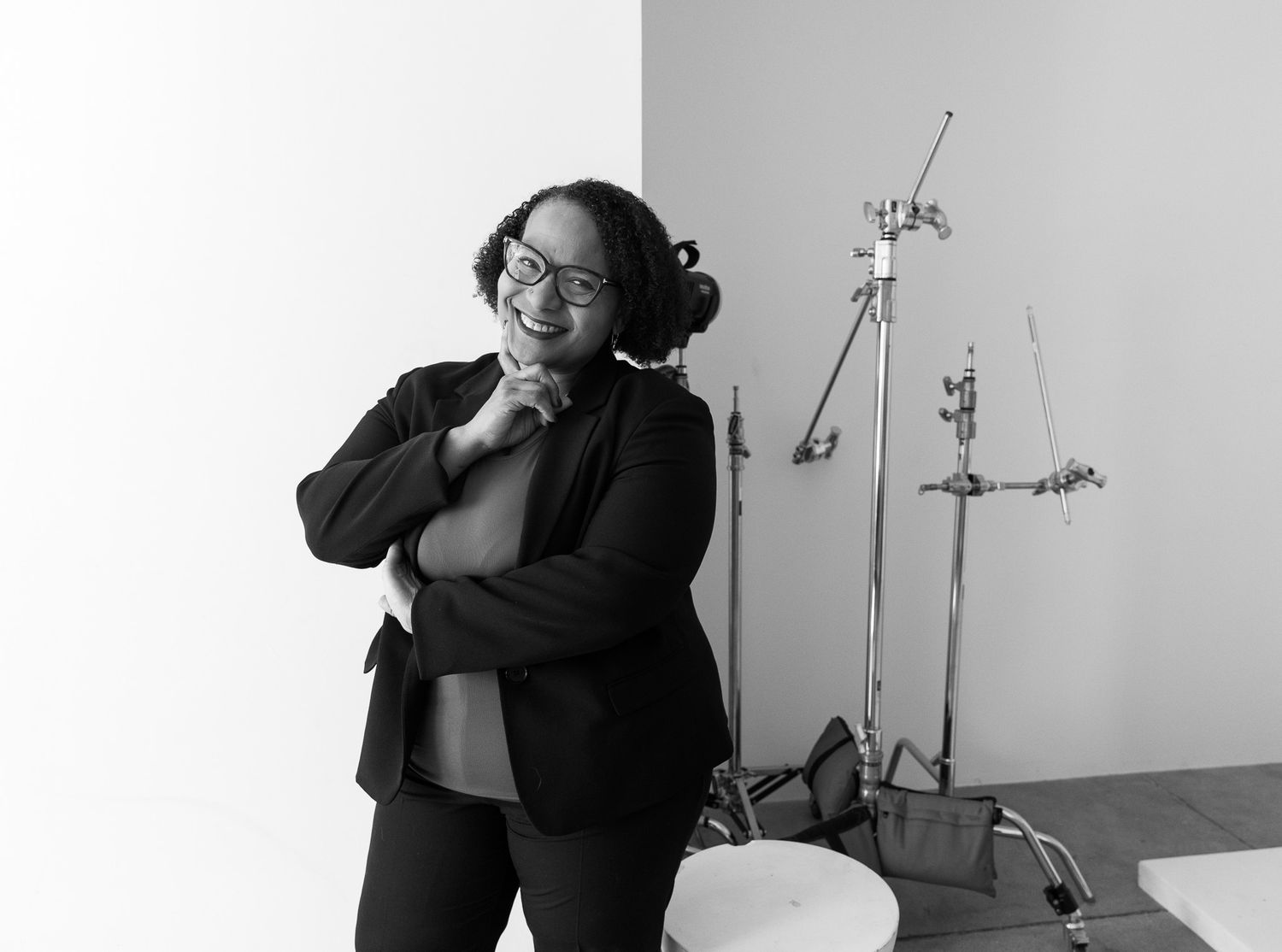 A smiling woman in glasses and blazer poses in a black and white photo studio setting.