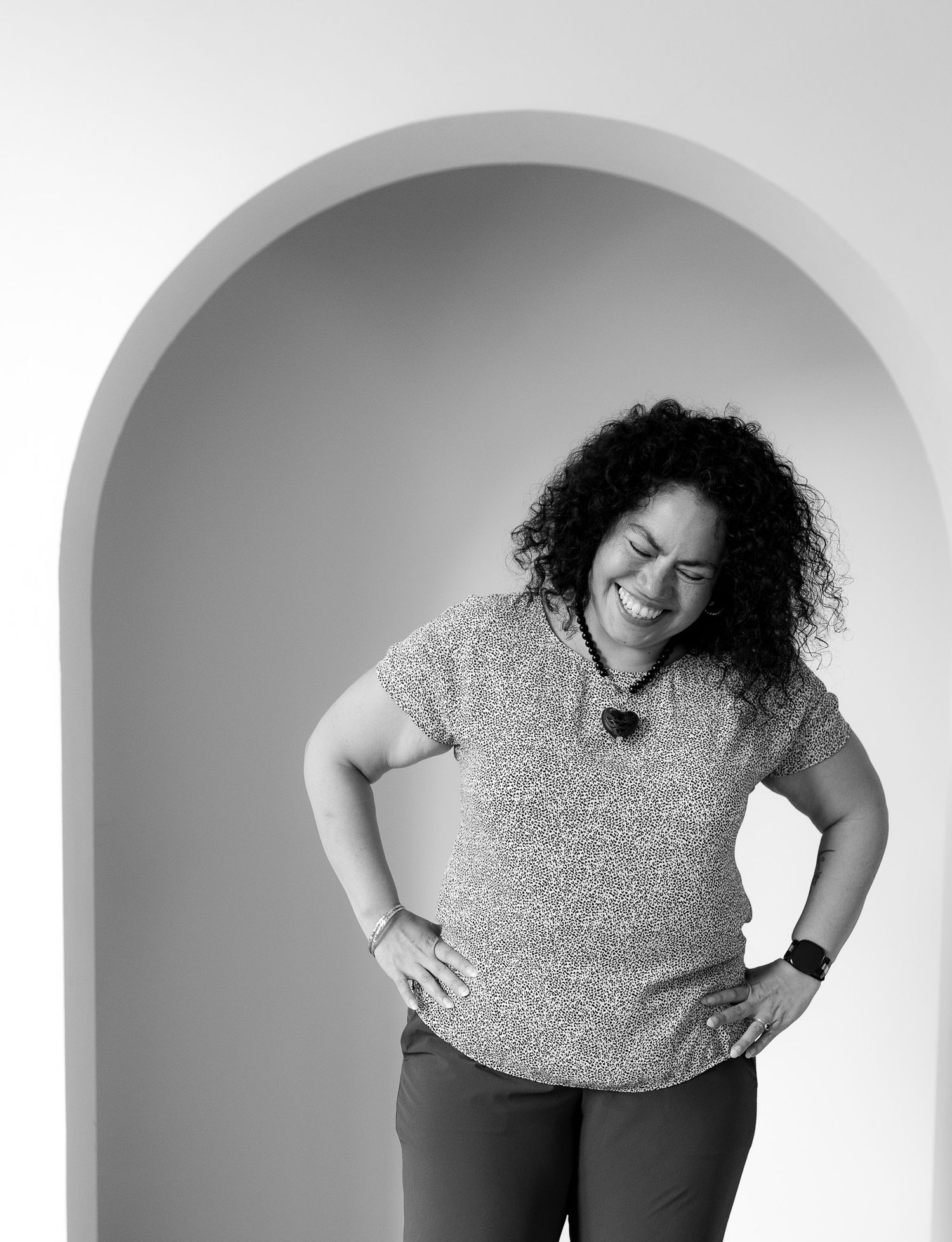 Black and white photo of a curly-haired woman smiling with hands on hips, standing in an arched doorway.