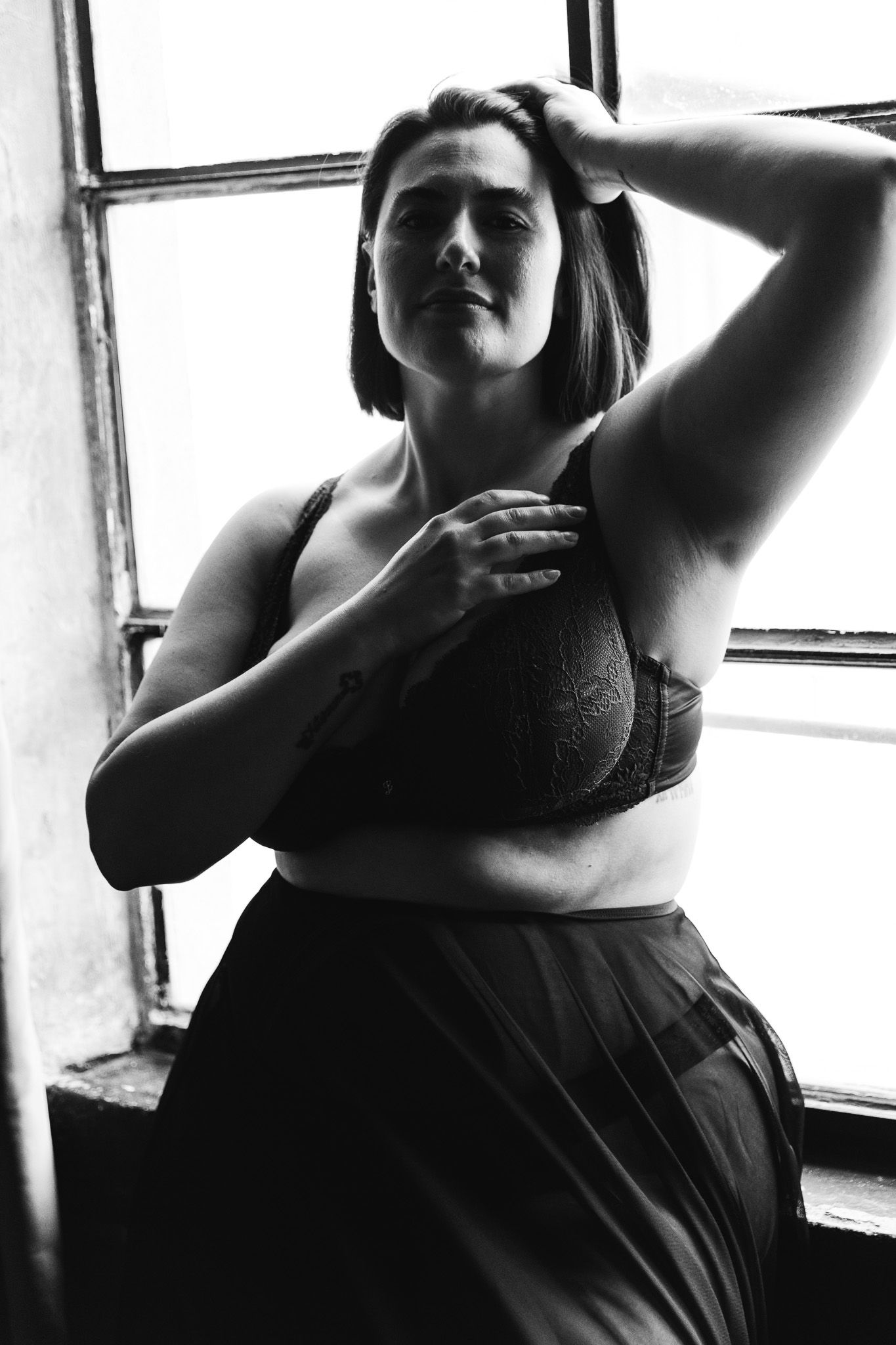 Black and white boudoir portrait of a woman in lace lingerie posing confidently by a bright window.