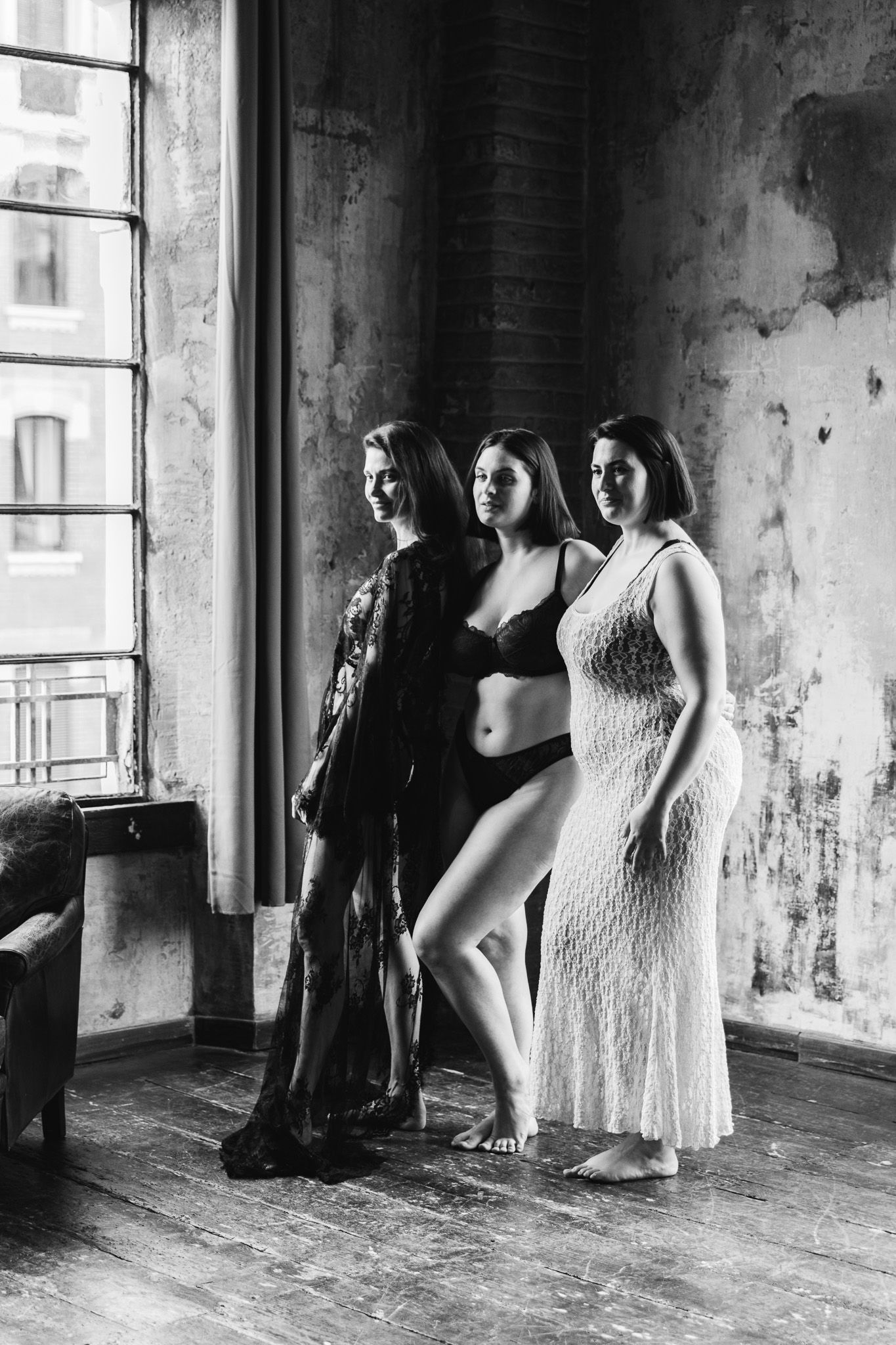 Three women pose in lingerie and sheer robes in a black and white boudoir photo shoot in a rustic loft.