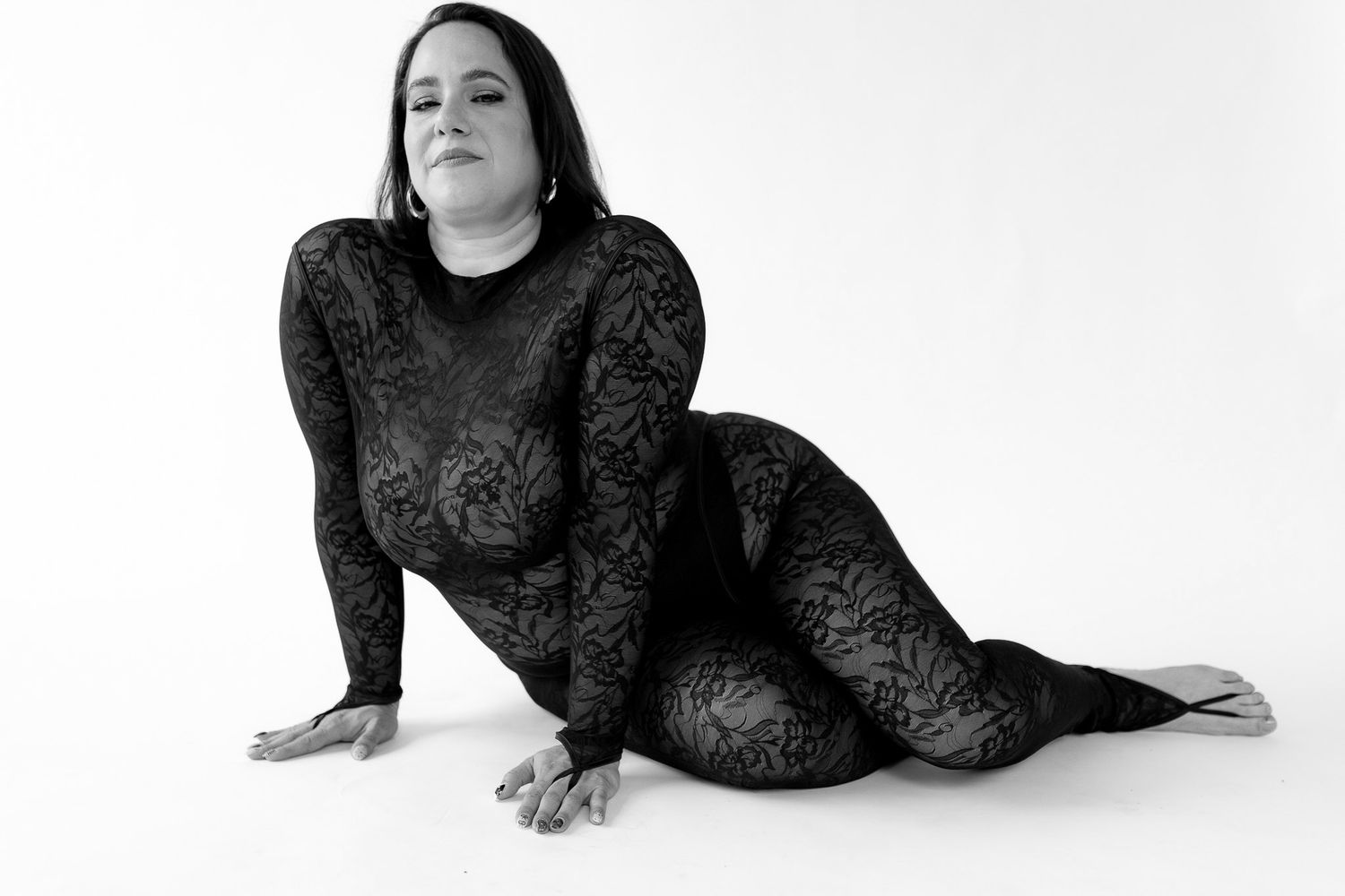 Woman in black lace bodysuit poses seated on floor in artistic black and white portrait.