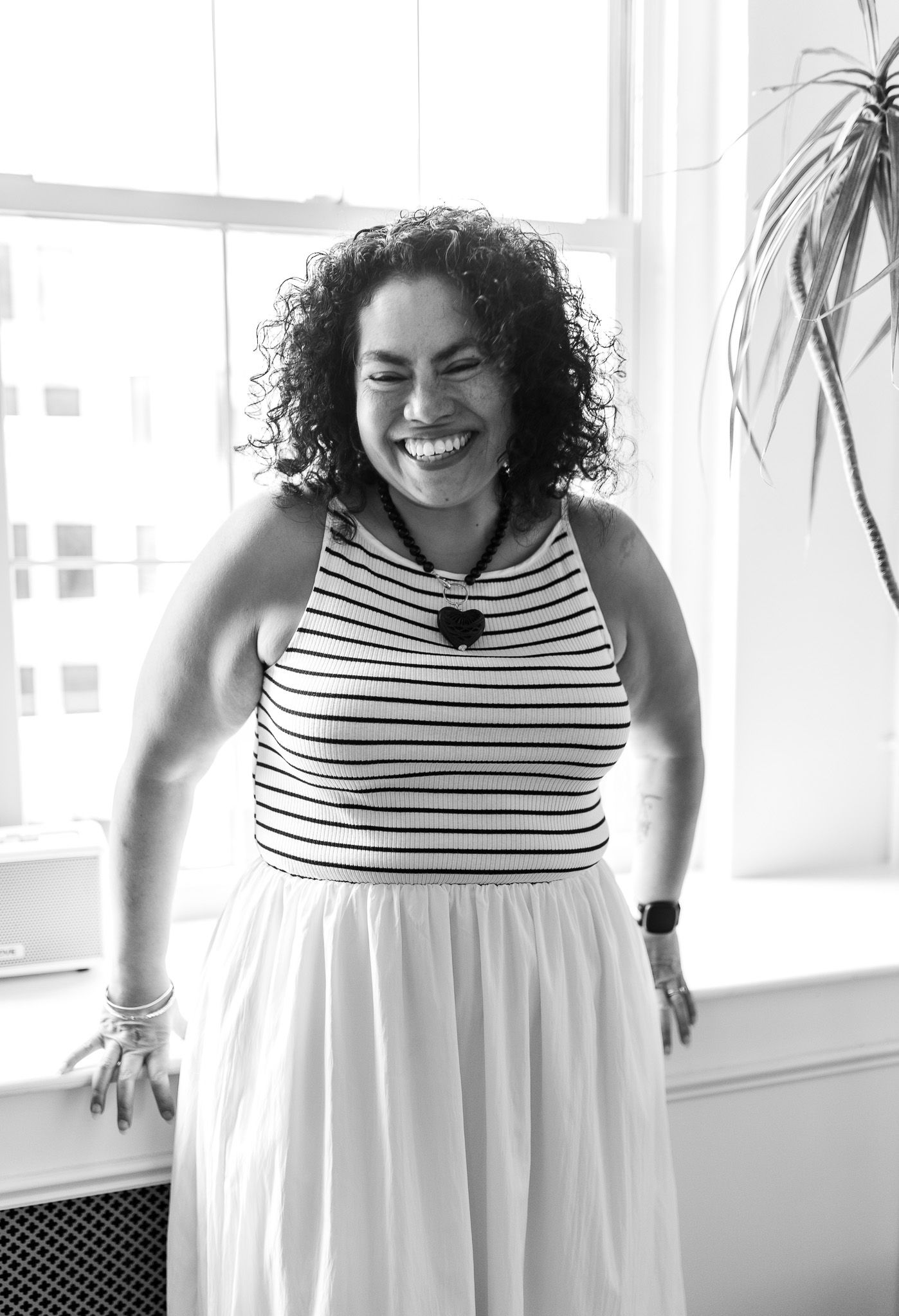 Woman with curly hair laughing joyfully, wearing striped top and white skirt, standing by bright window.