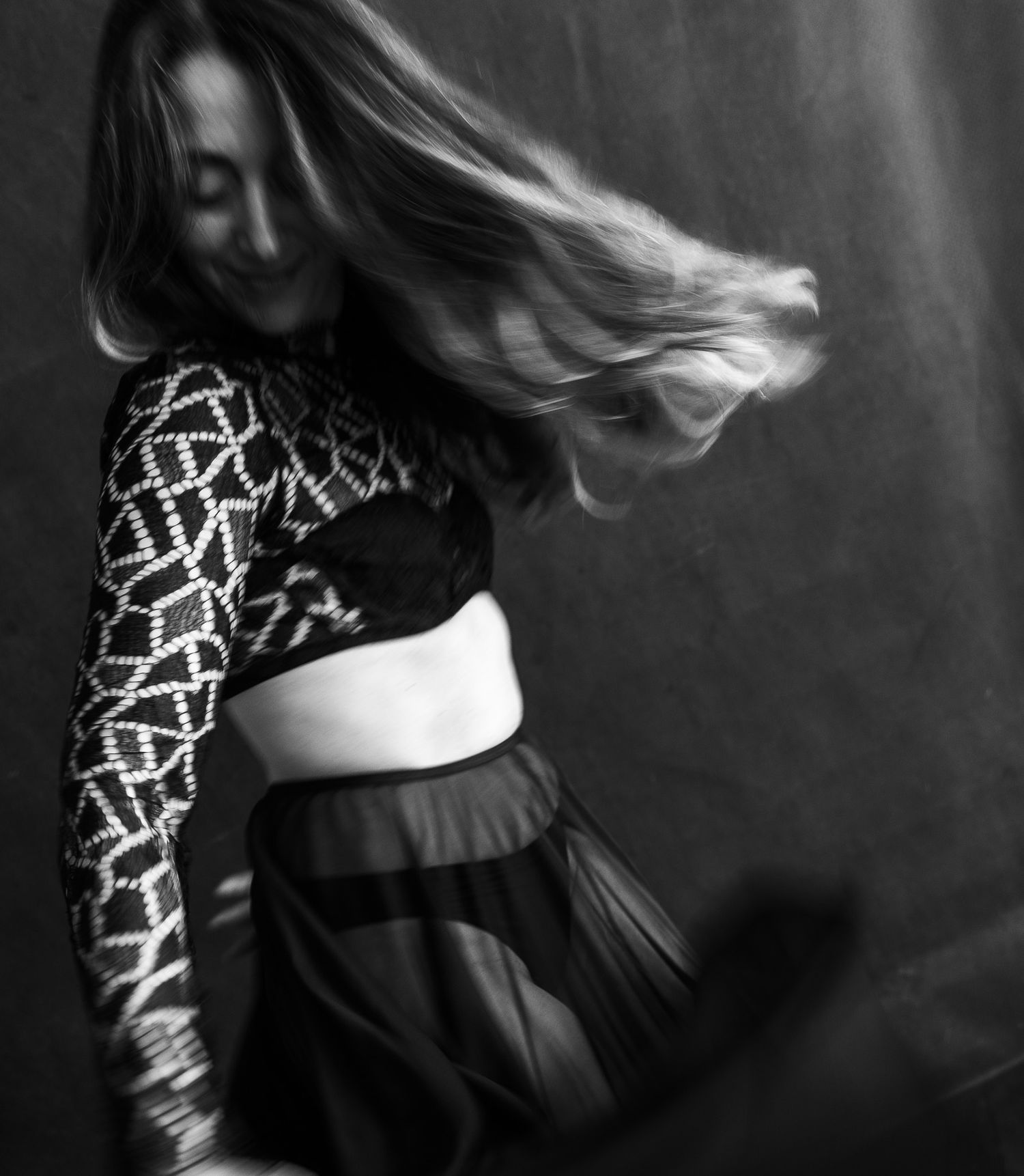Black and white photo of a woman dancing with hair in motion, wearing a patterned crop top and sheer skirt.