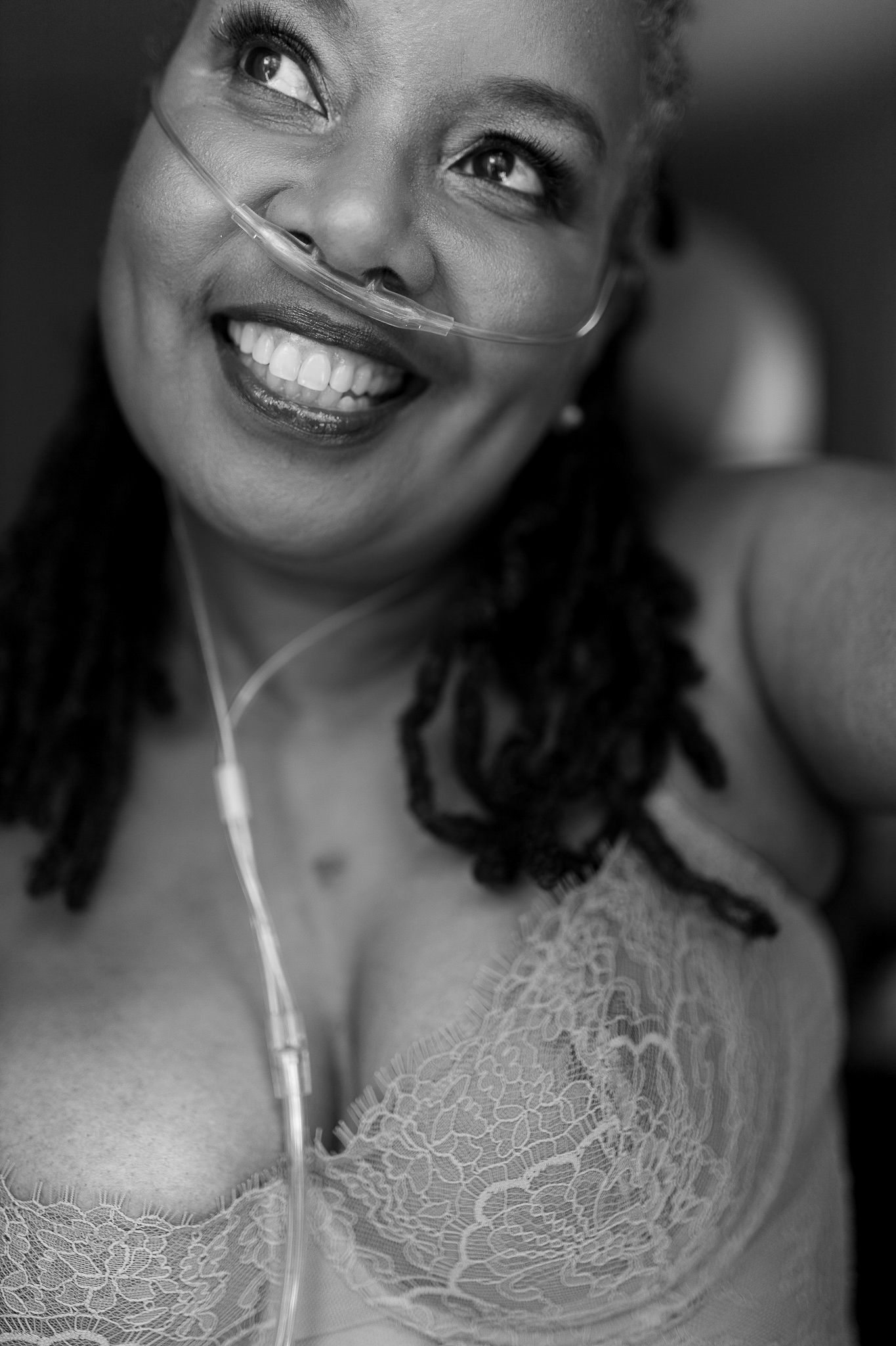Smiling Black woman wearing nasal cannula and lace bra looking upward in black and white portrait.