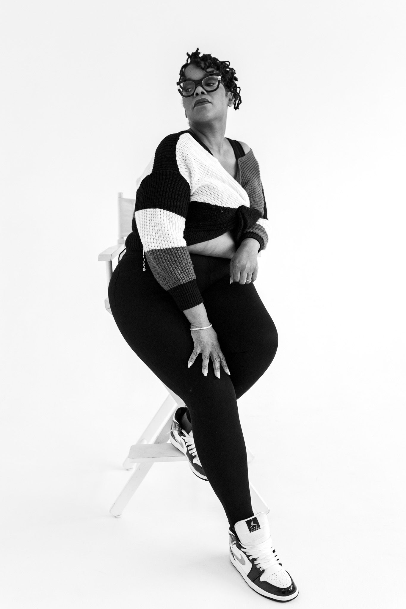 Black and white photo of a woman in glasses, striped sweater, black leggings, and Air Jordans sitting on a chair.