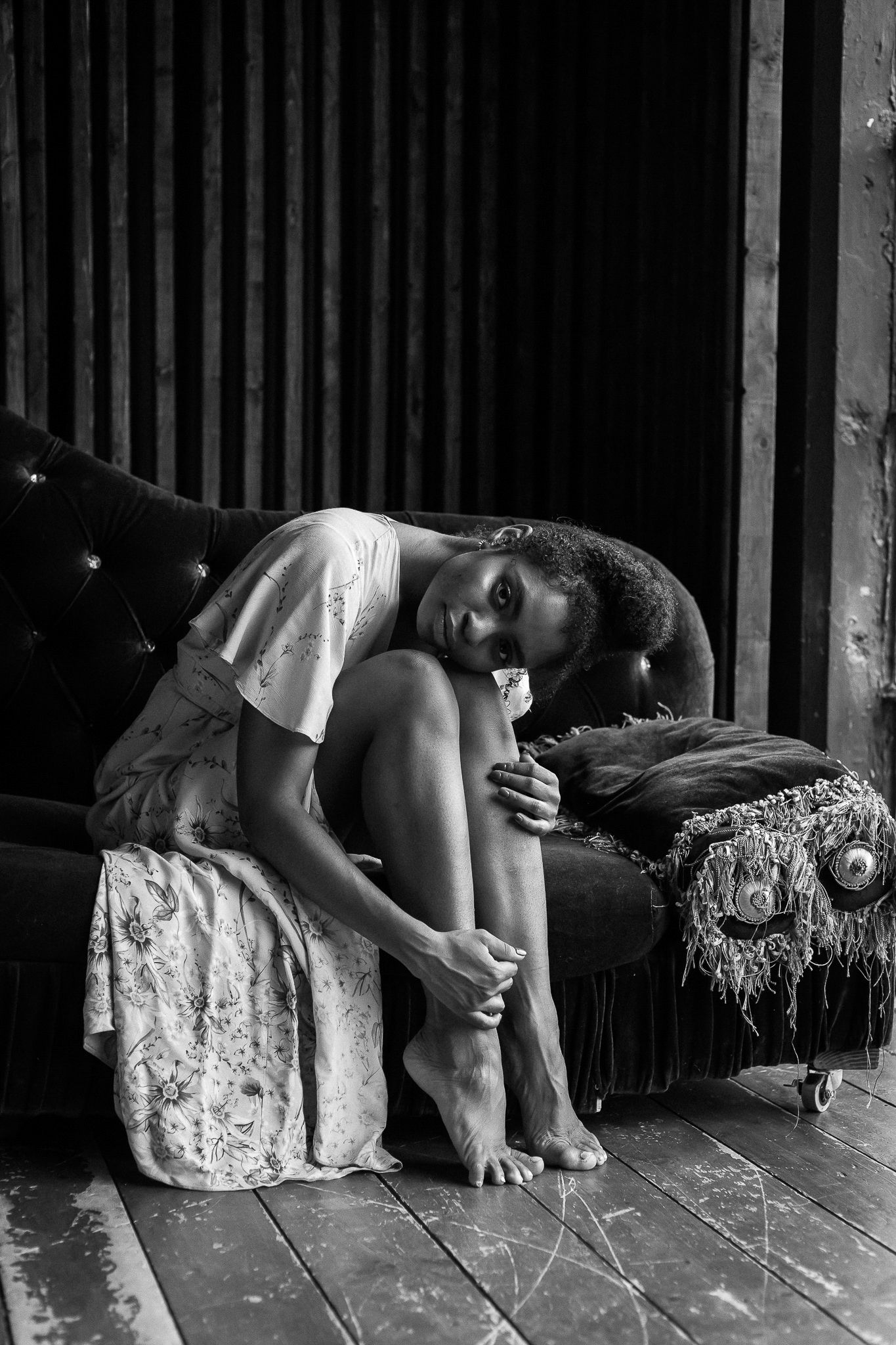 Black and white portrait of a young woman in a floral dress sitting on a vintage sofa.