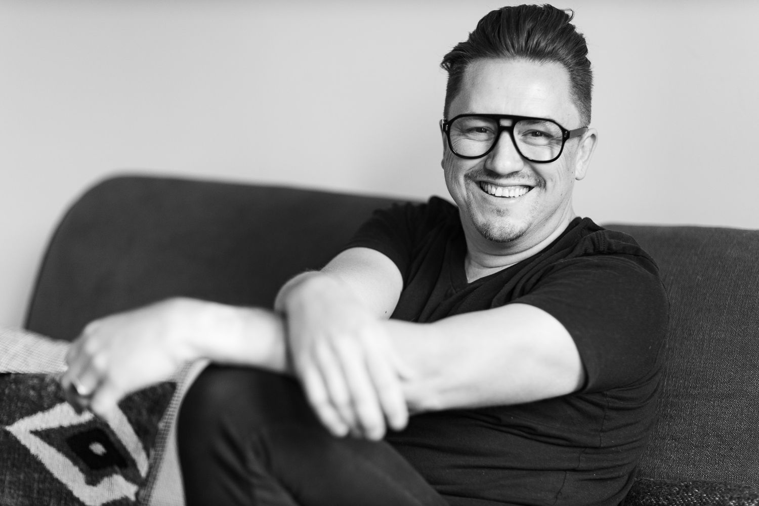 Man in black t-shirt and glasses smiling while relaxing on a couch.