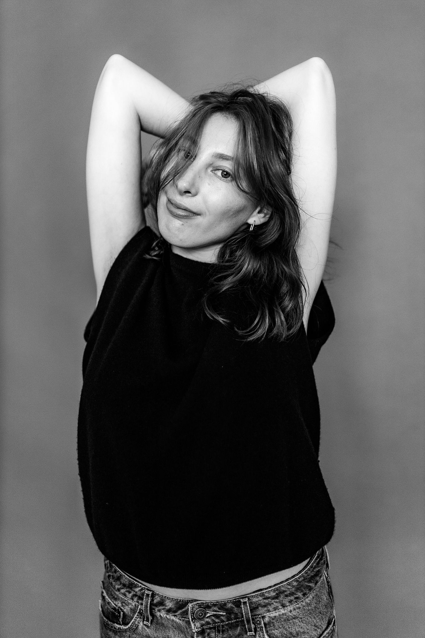 Black and white portrait of smiling woman in black crop tee with arms raised behind head in studio.