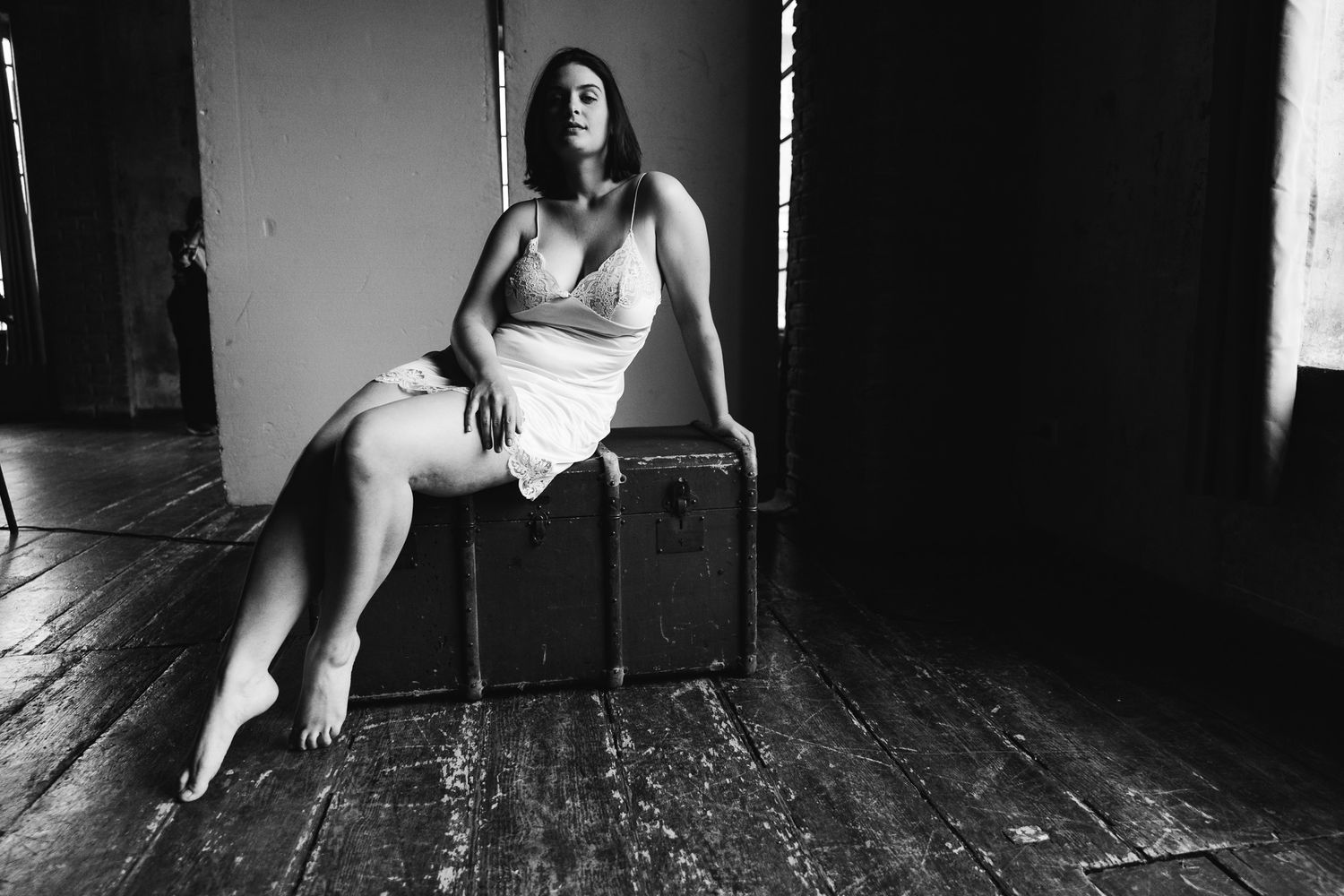 Black and white boudoir photo of a woman in a slip dress seated on an antique trunk.
