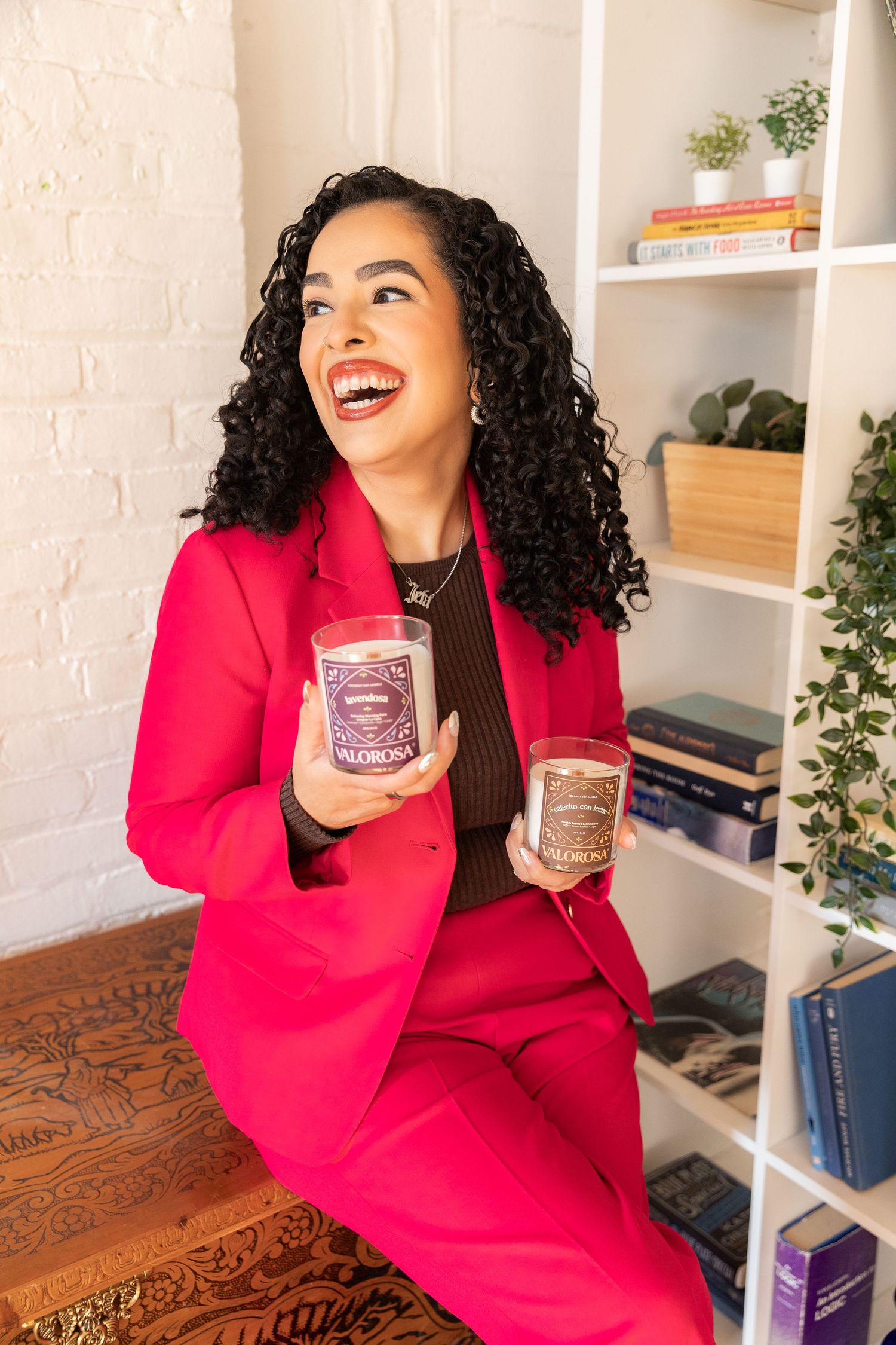 Smiling woman in pink blazer holding two decorative candles, sitting near a white bookshelf with plants and books.