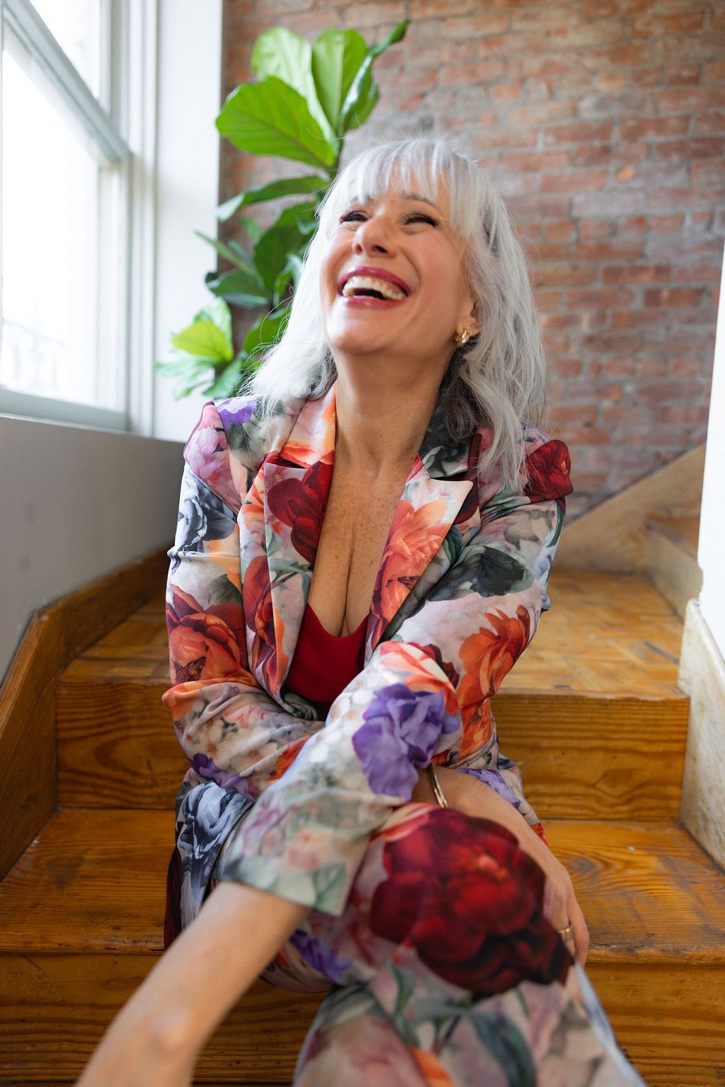 Laughing older woman with gray hair wearing a floral suit sits on wooden stairs near a brick wall and plant.