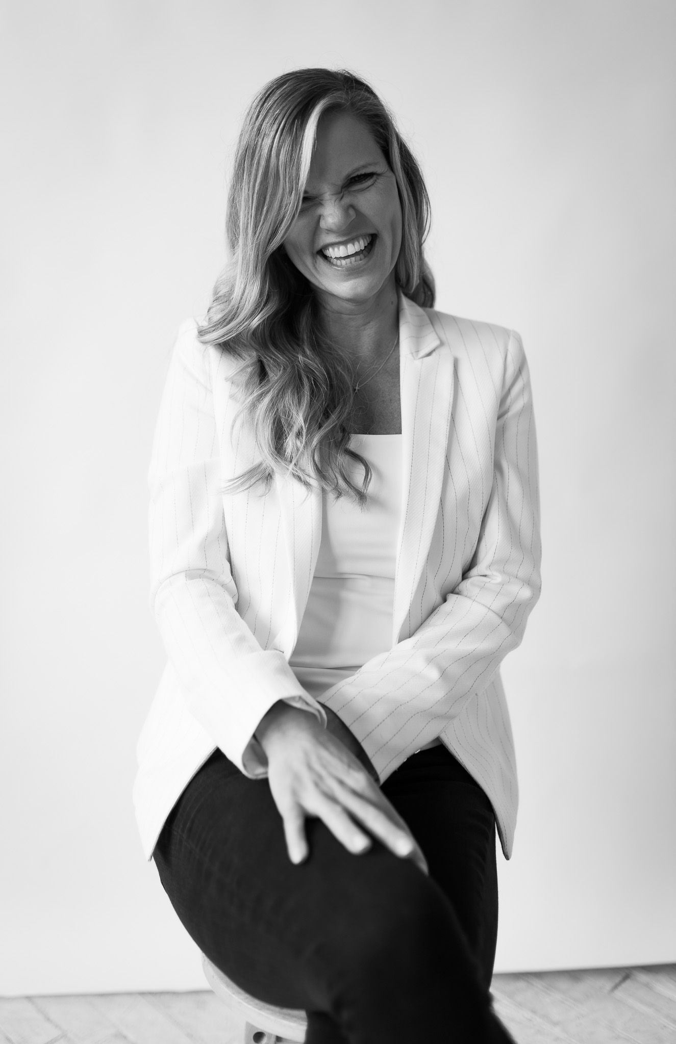 Blonde woman in white blazer laughing joyfully while seated, black and white studio portrait.