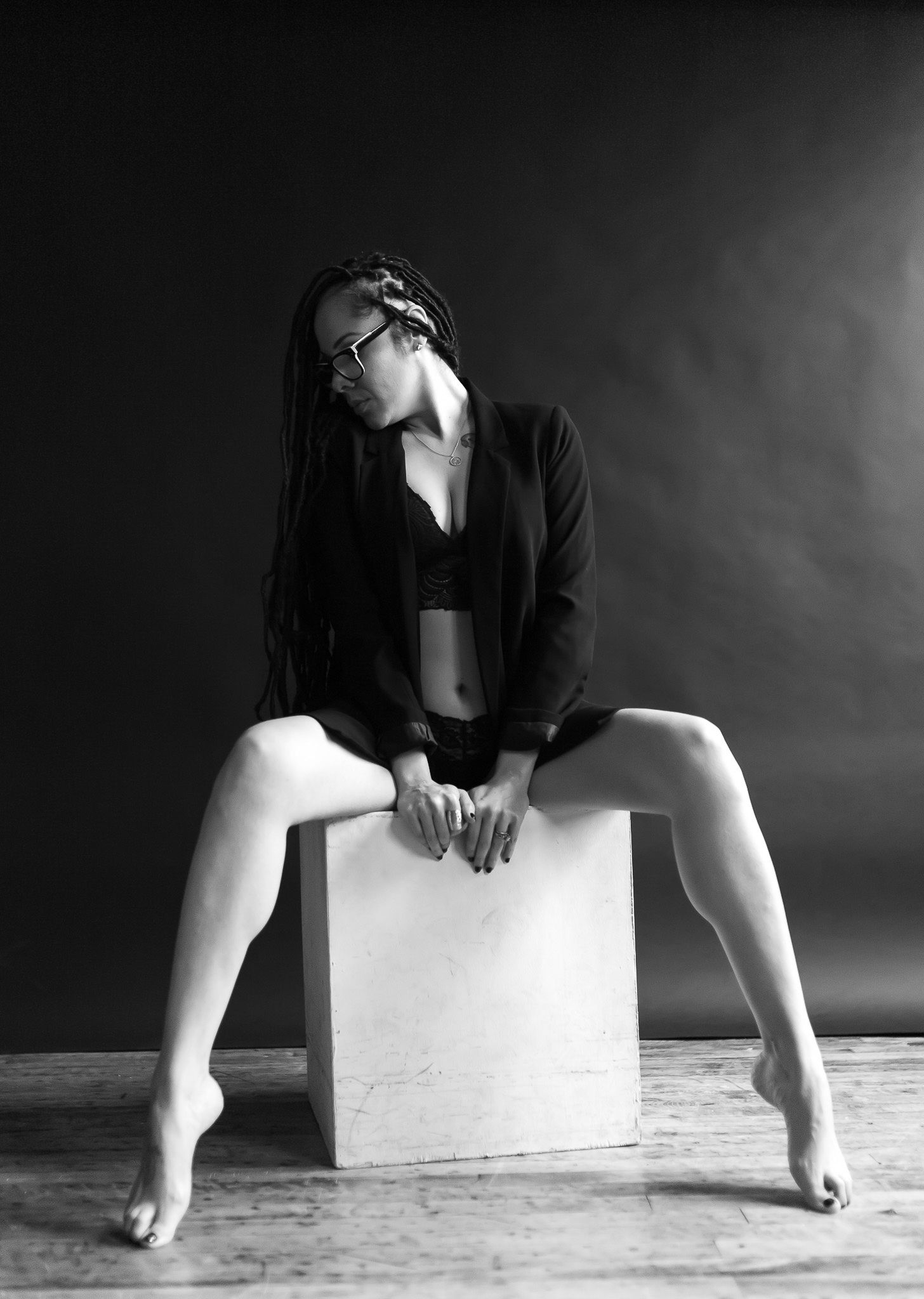 Black and white studio portrait of a woman with long braids and glasses sitting on a white cube in a black blazer.