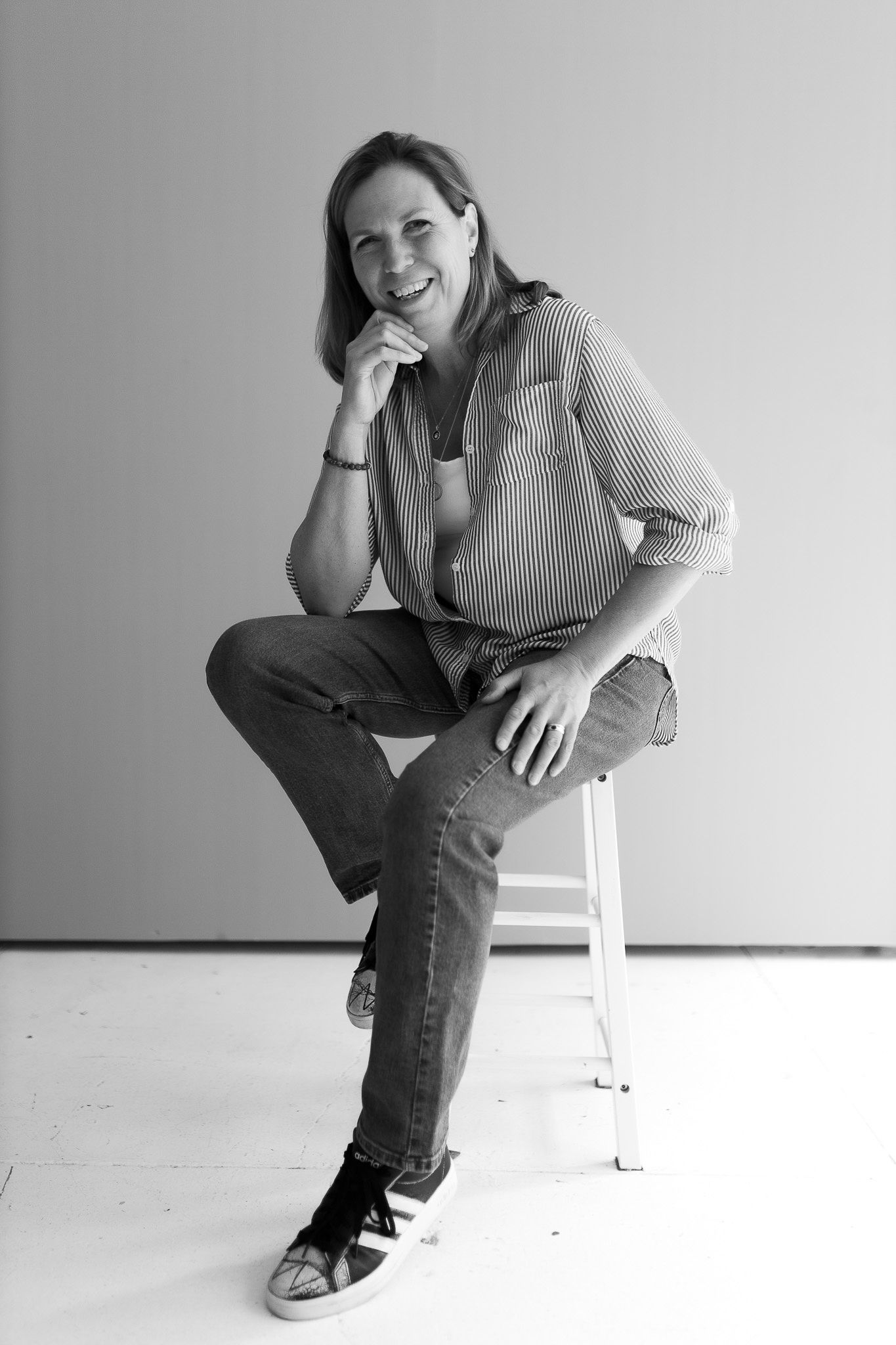 Black and white portrait of a relaxed woman sitting on a stool, smiling with hand on chin.
