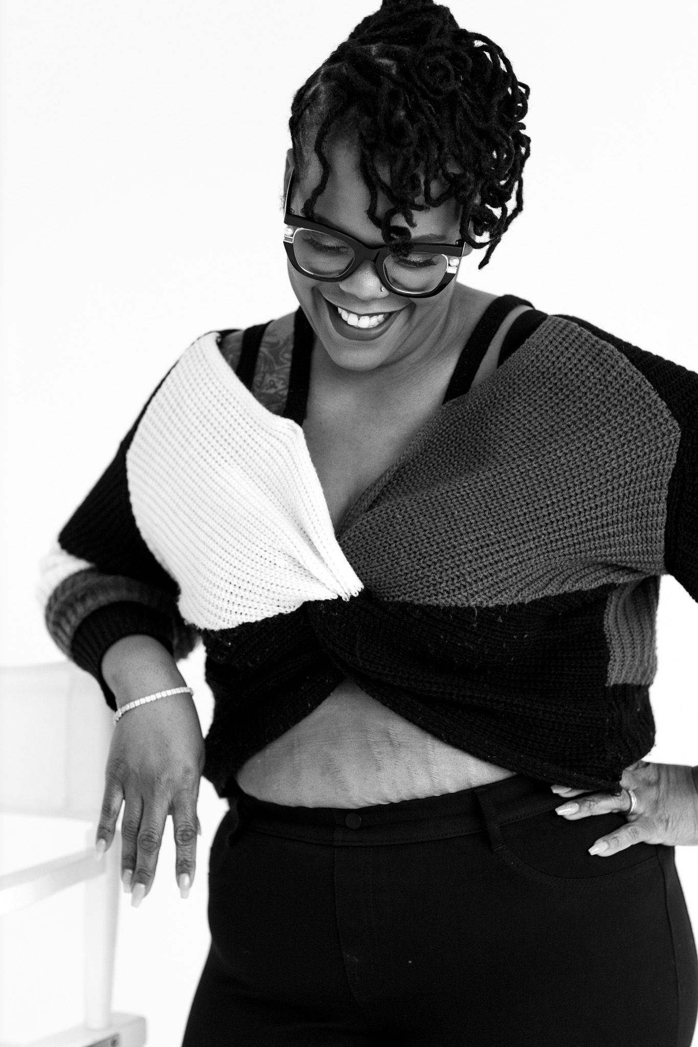 Black and white photo of a smiling woman in glasses wearing a colorblock knit sweater with midriff showing.