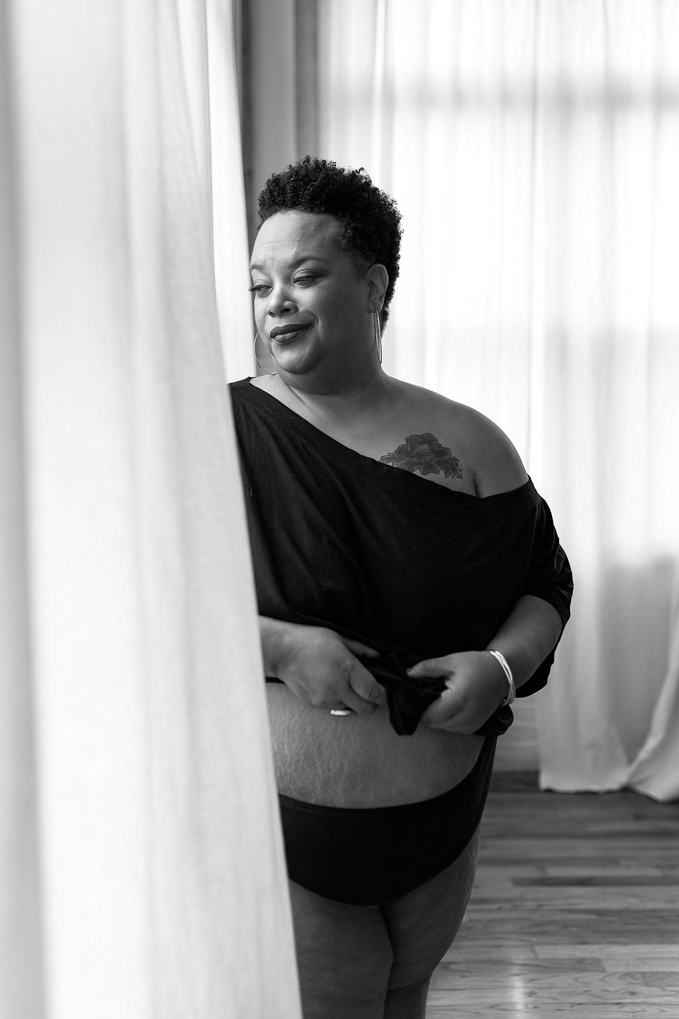 Black and white photo of a plus-size woman in a black off-shoulder top and shorts smiling by sheer curtains.