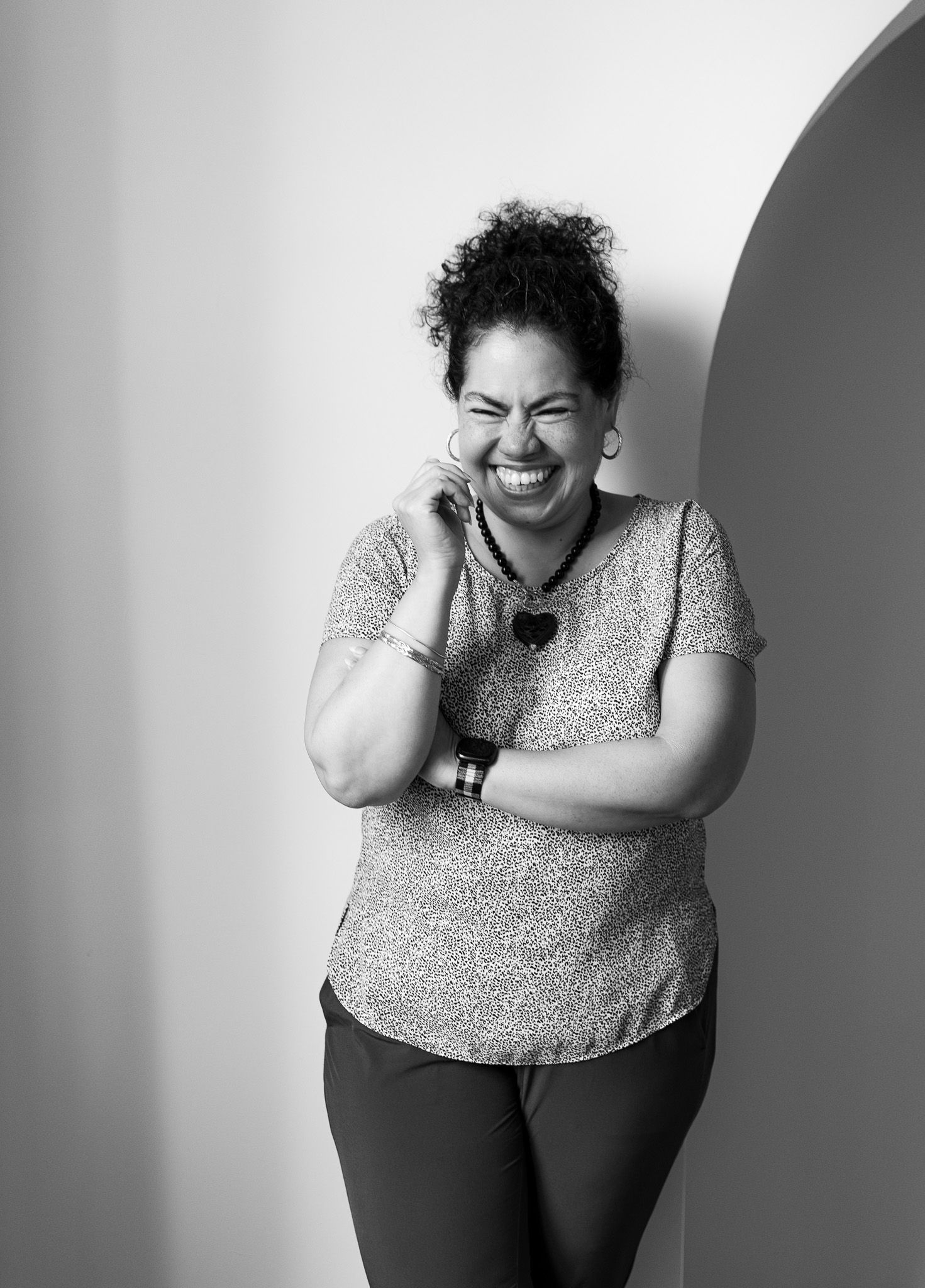 Black and white portrait of a joyful woman with curly hair laughing, leaning against a wall with an arch.