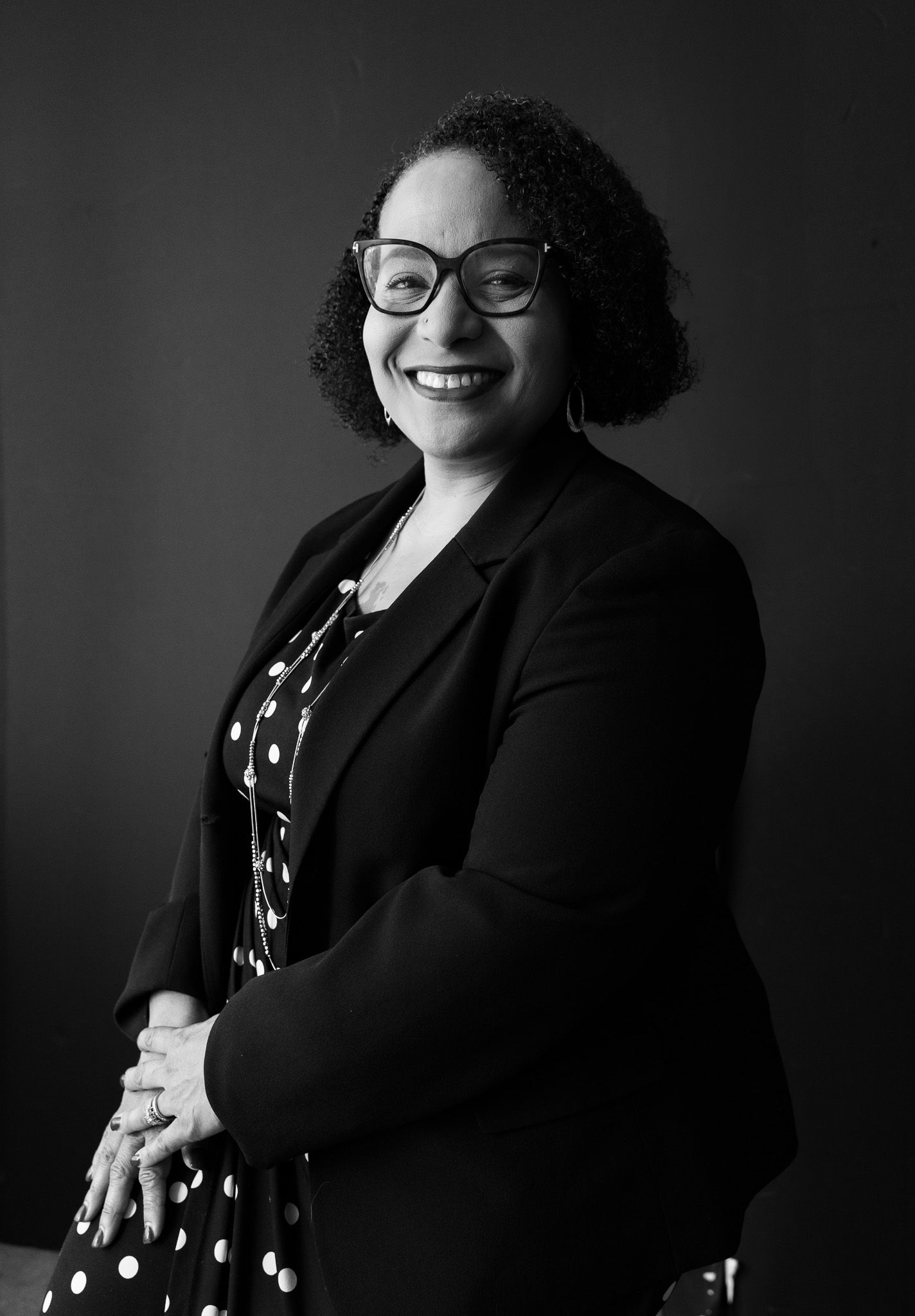 Professional black and white portrait of a smiling woman wearing glasses and a polka dot dress with blazer.
