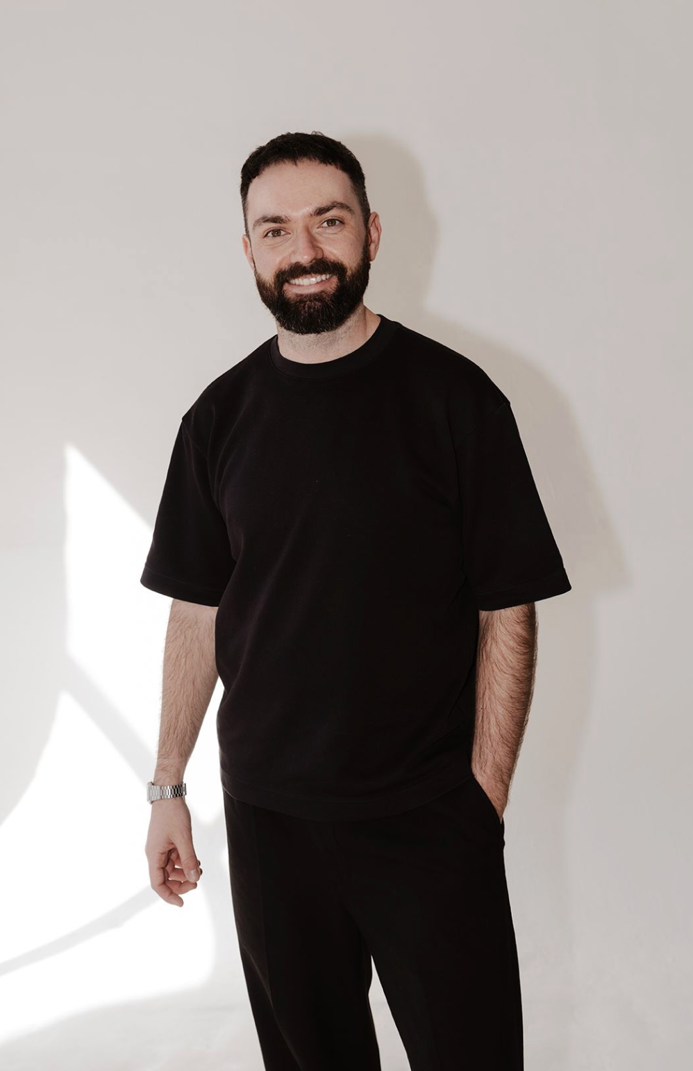 Smiling bearded man in black oversized t-shirt and trousers standing against white wall with natural light shadow.