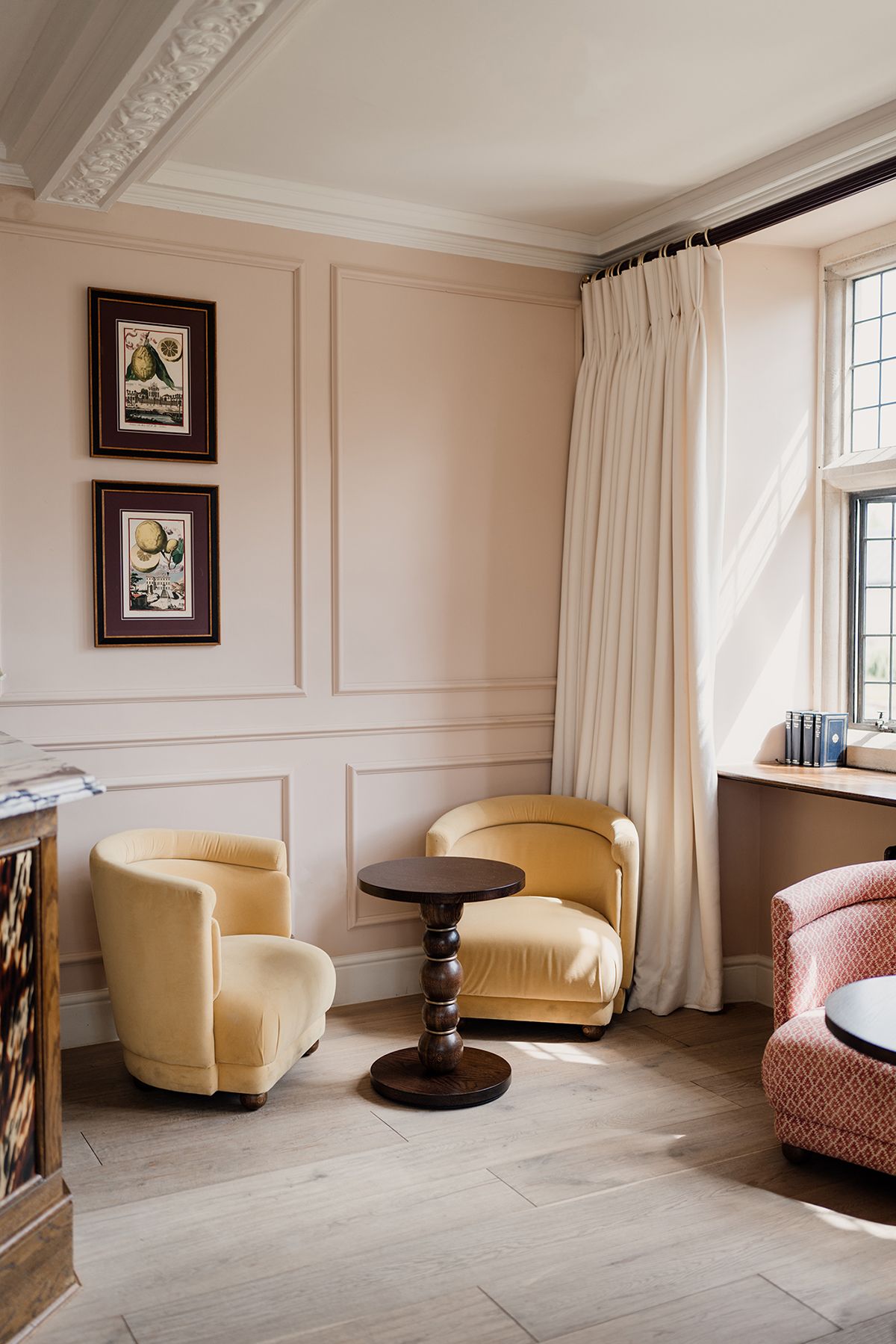 Elegant sitting room with pale pink walls, two yellow velvet armchairs, dark pedestal table, and tall cream curtains.