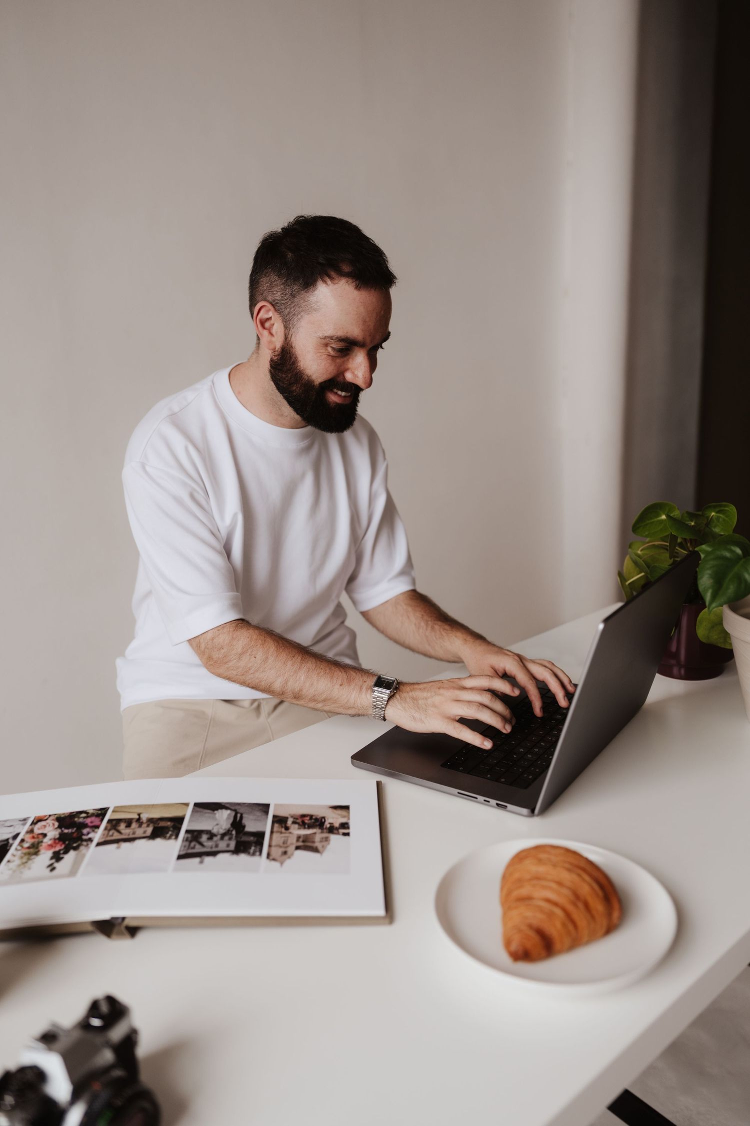 Smiling bearded man in white t-shirt working on laptop at white desk with croissant and photo album.