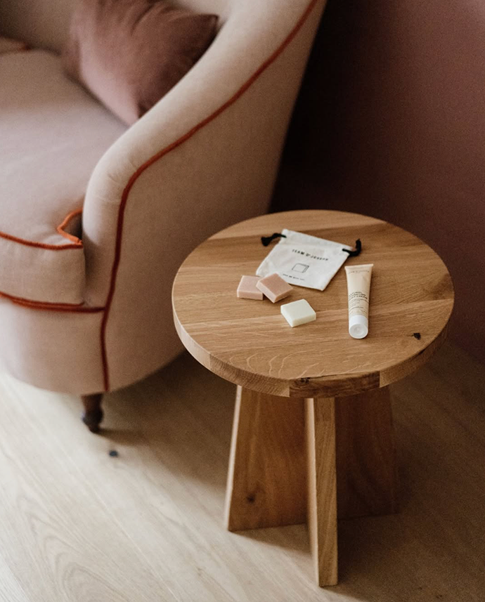 Round oak side table with toiletries and a small pouch beside a blush pink sofa on light wood floor.