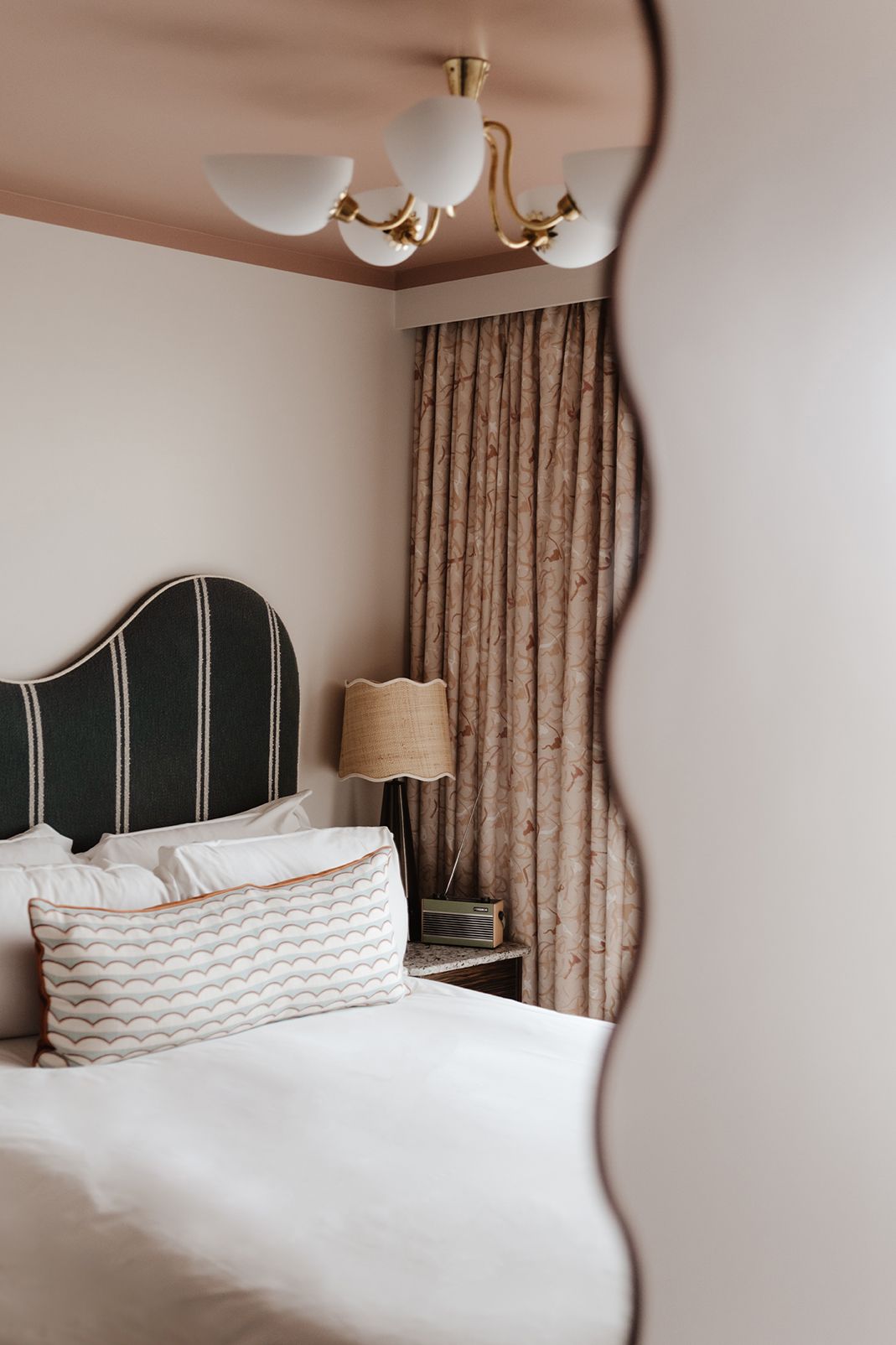Elegant hotel bedroom reflected in wavy mirror, featuring striped headboard, quilted pillows, and gold chandelier.