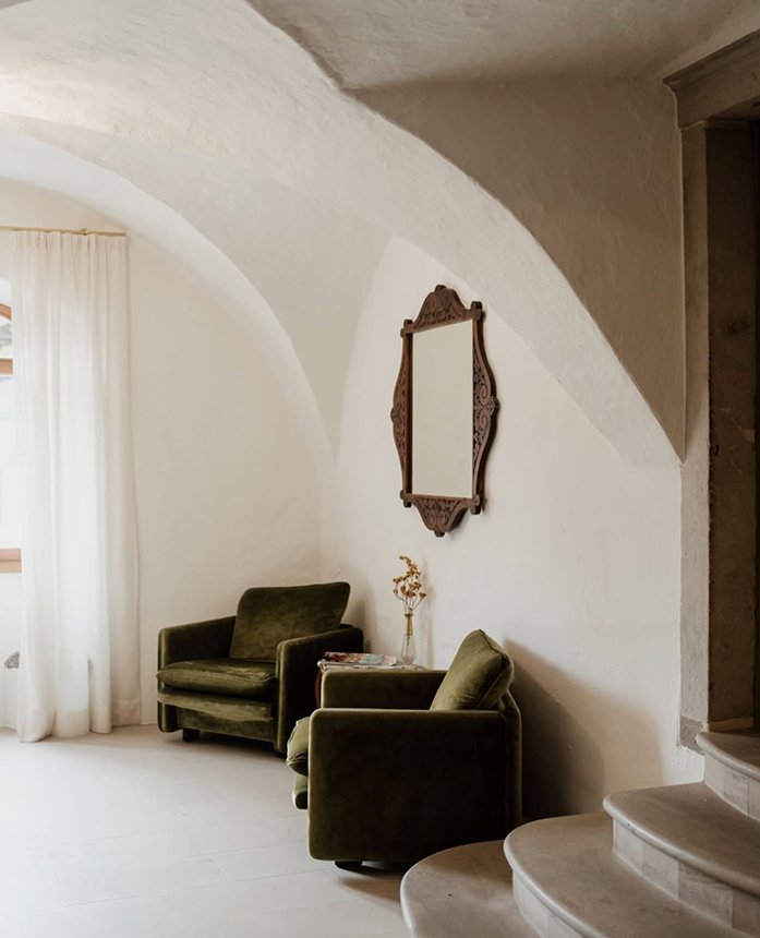 Rustic arched white plaster interior with green velvet armchairs, ornate mirror, and curved staircase.
