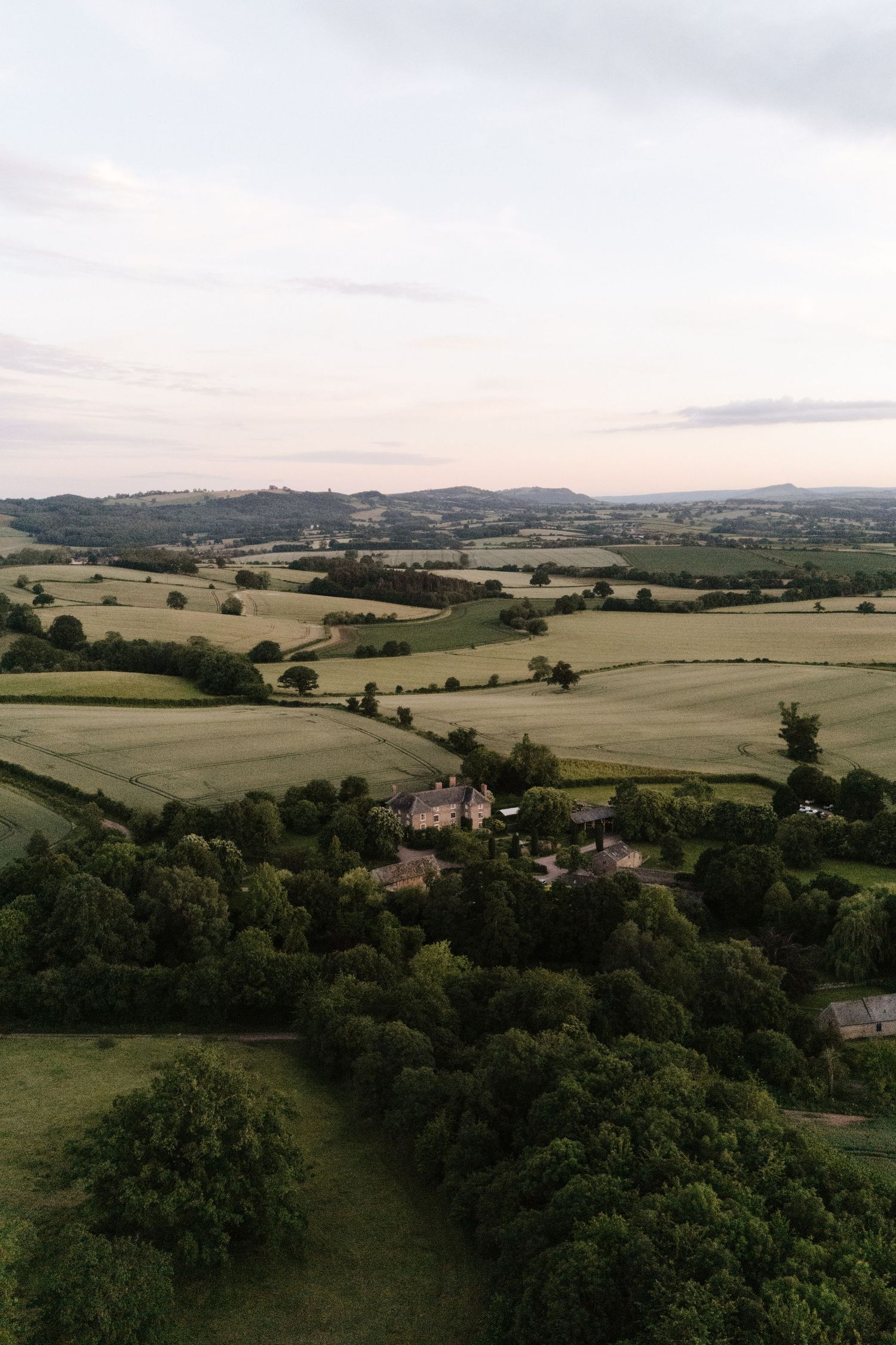 Aerial view of lush green English countryside with rolling fields, scattered trees, and a farmhouse nestled among woodland at dusk.