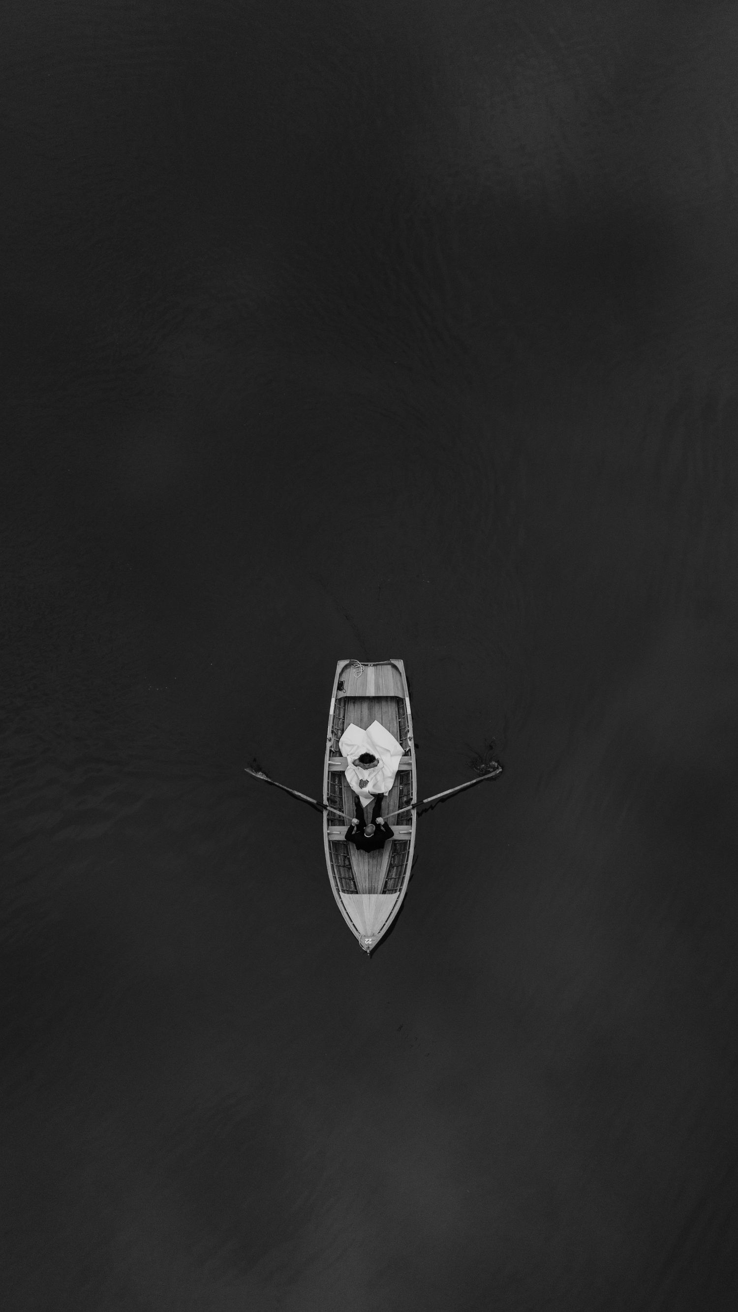 Aerial black and white view of a lone person rowing a small wooden boat on calm dark water.