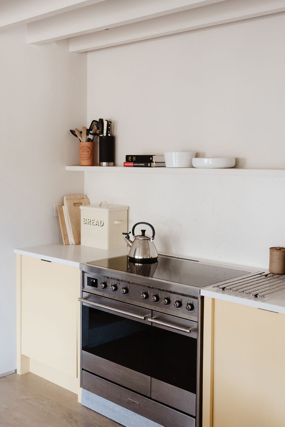 Minimalist kitchen with pale yellow cabinets, stainless steel range cooker, kettle, and open shelving with ceramics.
